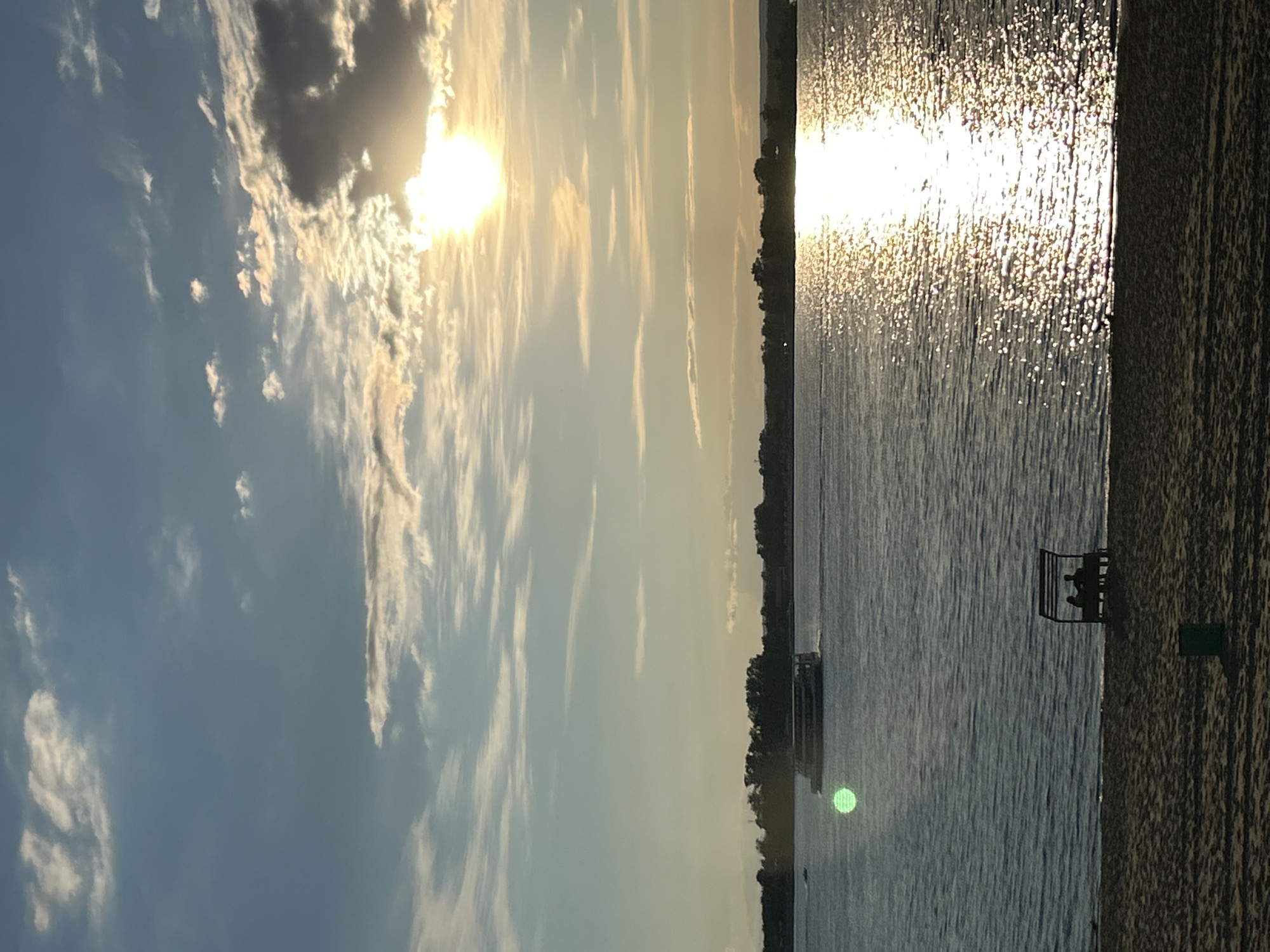 The image depicts a tranquil scene of a sunset over a body of water, likely the Volga River, near Novokuybyshevsk, Russia. In the foreground, a sandy beach is visible, with a small bench under a canopy occupied by two silhouetted figures. A green trash can is also present on the beach. The middle ground is dominated by the expanse of the water, reflecting the golden hues of the setting sun. A ferry boat is seen cruising in the distance. The background features a treeline along the distant shore. The sky is a canvas of soft blues and whites, with scattered clouds illuminated by the warm glow of the setting sun.
