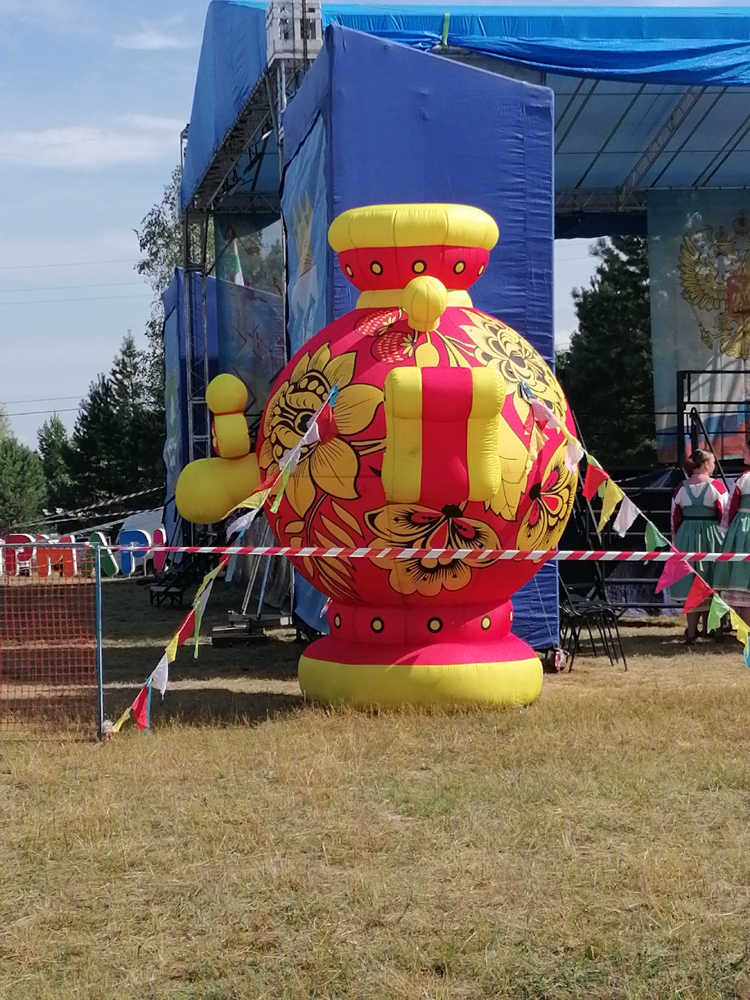 An outdoor festival scene unfolds under a clear blue sky with scattered clouds. Dominating the foreground is a large, inflatable, red and yellow object decorated with floral patterns. This object appears to be a stylized representation of a traditional Russian matryoshka doll or a samovar. Small, colorful flags on strings are strung across the area in front of the inflatable.
Behind the inflatable, a blue stage structure is visible, partially covered with blue tarps. Metal scaffolding supports the stage, and a large banner with an image of the Russian double-headed eagle crest is displayed on the stage backdrop.
To the right of the inflatable, two women are partially visible, wearing traditional, light green dresses with embroidered bodices and white blouses. They are standing behind a barrier, looking towards something off-camera.
In the middle ground, a red mesh fence is present on the left side of the image. Several folding chairs are positioned in front of the stage. The ground is covered in dry, mowed grass. The overall impression is of a festive outdoor event in a rural or park-like setting.
While the image depicts a scene that could be from a festival, there is no specific visual information within the image that allows for pinpointing the exact location to the city of Shadrinsk, Russia. The presence of the Russian eagle crest suggests a Russian context, but not a specific city.