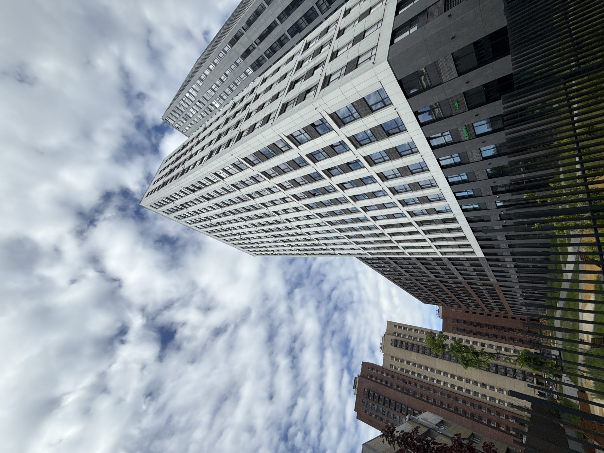 Looking up at several high-rise buildings in Moscow, Russia, against a cloudy sky. Buildings vary in color, including white, gray, and brick red. Metal bars are visible in the foreground, possibly a fence.
