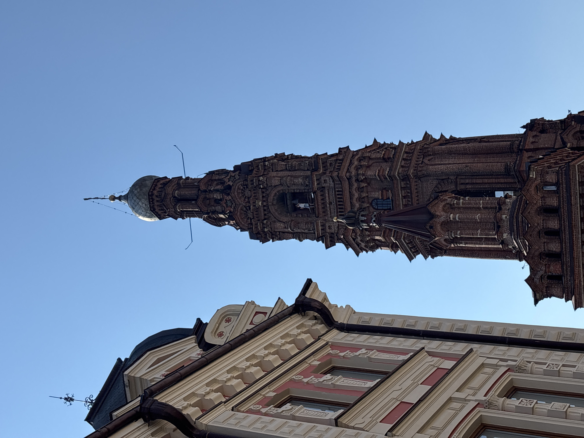 The image shows a view of the exterior of the Cathedral of the Annunciation in Kazan, Russia, on a clear day. The foreground features the detailed facade of a building with ornate carvings and painted accents, partially obscuring a tall, dark brick bell tower. The bell tower, which dominates the right side of the frame, is intricately designed with multiple tiers and arched openings. A person can be seen standing in one of the open arches of the tower. The sky is a vibrant blue.