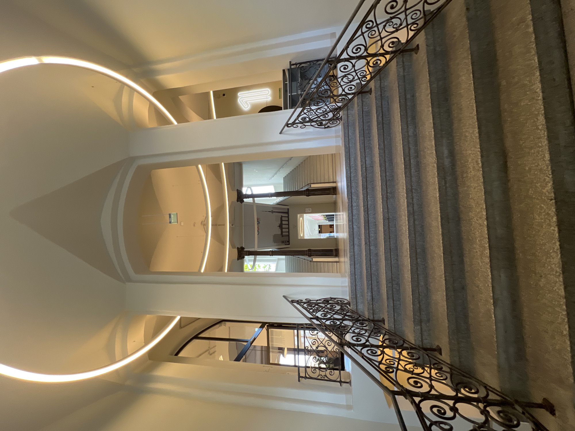 The image displays the interior of a grand building, rotated 90 degrees clockwise. On the right, a wide, light-colored staircase ascends, featuring numerous steps that cast distinct shadows. An ornate, dark wrought-iron railing with intricate scrollwork runs alongside the staircase.
The left side of the frame is dominated by smooth, light-colored arched ceilings and walls. Warm, indirect strip lighting is integrated into these architectural curves, illuminating the space and highlighting its clean lines.
Looking deeper into the building, past a series of arches, further interior sections are visible. Large, fluted columns stand prominently in the mid-ground. On an upper level, a bright white neon sign in a stylized letter shape, possibly an "L" or "F," glows against a light wall. Below this level, through an opening or large window, another part of the building's interior can be seen.
In the distant background, a wall is visible with white text that reads "Google" and an outlined graphic of a city landmark, which appears to be the Brandenburg Gate. This detail, combined with the location context of Berlin, suggests the setting is likely a Google office or a related facility in Berlin.
No people are present in the image. The time of day is not discernible due to the reliance on interior lighting. The overall aesthetic combines classic architectural elements, such as the grand staircase and columns, with modern design, exemplified by the integrated strip lighting and contemporary signage.