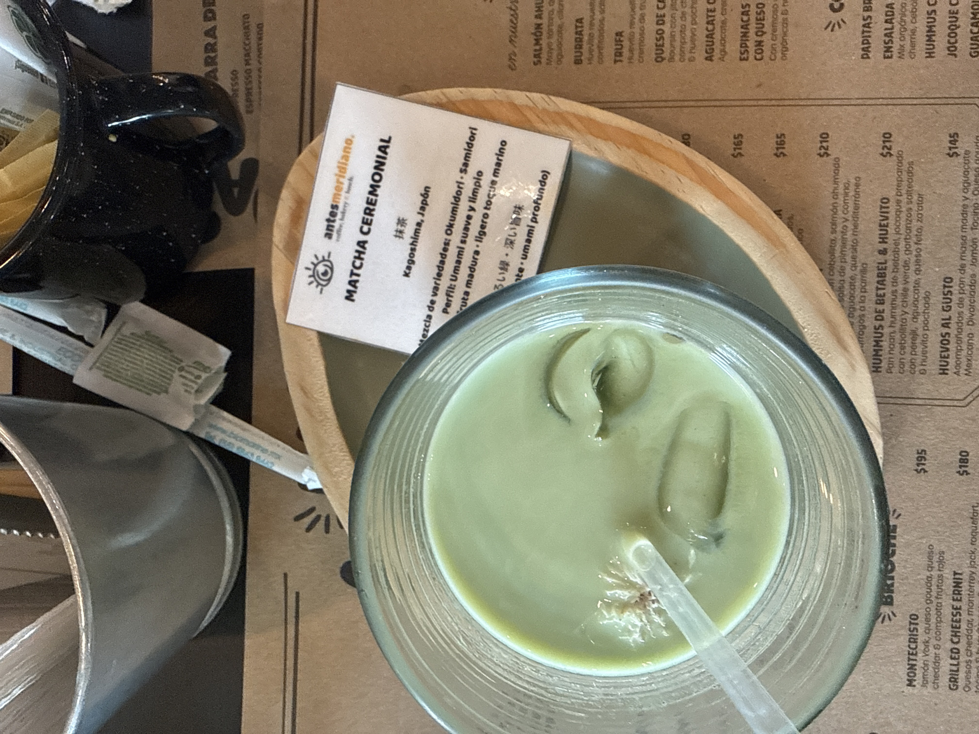 A high-angle shot depicts a Matcha Ceremonial drink in a glass, featuring ice cubes and a straw. Next to it is a menu card from "antesmeridiano. coffee, bakery & foods," describing the drink's characteristics in both Spanish and Japanese. The menu lists other food items and prices in pesos. Nearby are a black speckled mug, sugar packets, a silver metal container, and a wooden tray. The scene is located in Puebla, Mexico, based on the menu and branding.