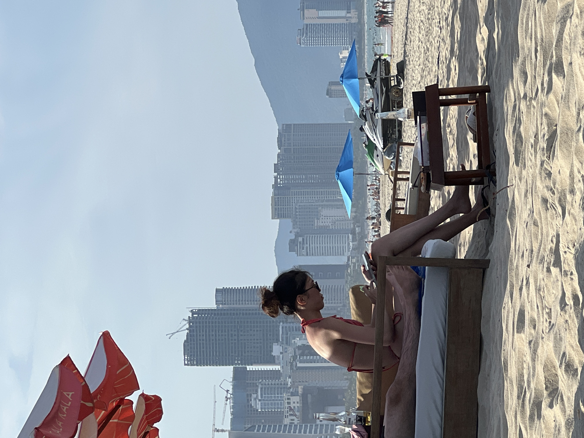 The image depicts a beach scene in Da Nang, Vietnam, with a woman in a red bikini sitting on a lounge chair and looking at her phone. She is positioned on a sandy beach with a low wooden table to her right. In the background, there are several high-rise buildings under construction and some completed skyscrapers, indicating an urban coastal environment. A large red and white umbrella is visible in the upper left corner. Further in the background, the ocean is visible with some people and jet skis on the water. The overall atmosphere is bright and sunny, suggesting a typical day at the beach.