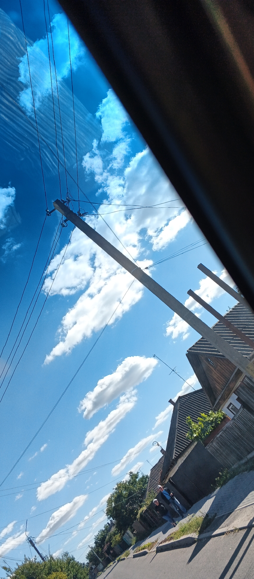 The image depicts a street scene in Smila, Ukraine, on a sunny day with a blue sky dotted with white clouds. The perspective is looking upwards and diagonally from a vehicle, indicated by the dark interior frame in the upper right corner and the windshield's reflection causing horizontal lines across the sky.
In the foreground, a paved road runs diagonally from the bottom left towards the bottom right. A sidewalk with some grass and foliage runs along the right side of the road. Two individuals are visible on the sidewalk, walking away from the camera. They are both wearing light-colored hats and dark jackets.
Further down the street on the right, there are buildings with wooden siding and corrugated roofs, typical of residential structures. A fence made of corrugated metal runs along the property line. Green trees and bushes are present, bordering the street and properties.
Overhead, a wooden utility pole with multiple wires is visible, angled across the sky from left to right. These wires stretch across the frame, adding a sense of the urban or suburban environment. The overall impression is a snapshot of everyday life in a Ukrainian town on a clear day.