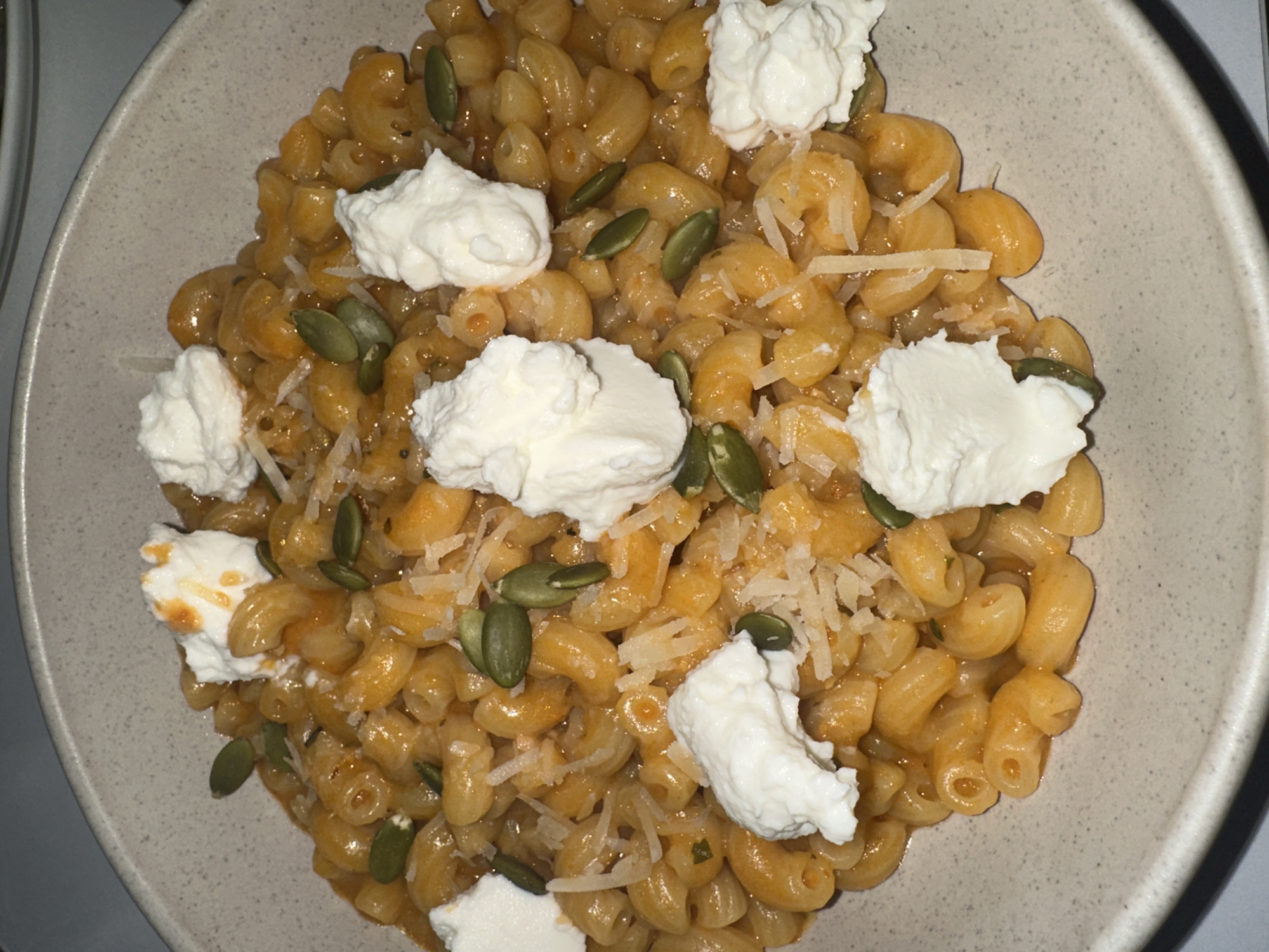 A close-up, overhead view depicts a single serving of pasta within a light gray, speckled ceramic bowl. The dish consists of small, curved macaroni-style pasta coated in a glossy, orange-red sauce. The pasta is topped with approximately eight distinct dollops of white, creamy cheese, some exhibiting faint orange drizzles. Numerous dark green, flat, oval-shaped pumpkin seeds are scattered across the surface, interspersed with a sparse quantity of finely shredded white or pale yellow cheese. The bowl rests on an unidentifiable light-colored surface. The background is dark and out of focus. No individuals, actions, or specific environmental identifiers are visible to determine a precise location in Yerevan, Armenia.