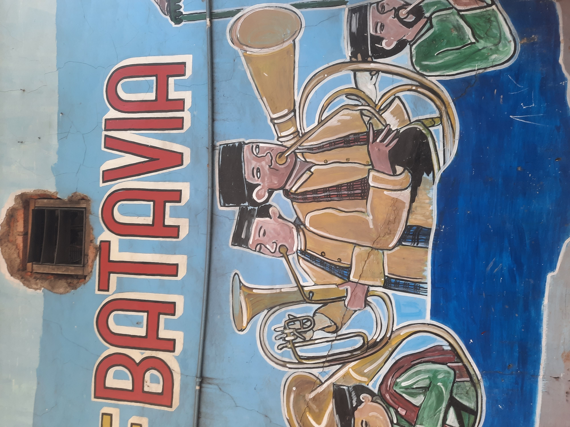 The image is a mural on a light blue, weathered wall depicting a brass band. The mural features the word "BATAVIA" in large red and white lettering across the top. Below the lettering, four men are depicted playing various brass instruments. The men are wearing traditional attire, including peci hats and checkered scarves. The mural is colorful, with yellow instruments and brown, green, and red clothing. The background is a vibrant blue, with some cracks and imperfections visible in the wall. The lower portion of the mural shows a blue wave-like pattern. The mural is likely located in North Jakarta, Indonesia, given the historical significance of Batavia as the former name of the city.