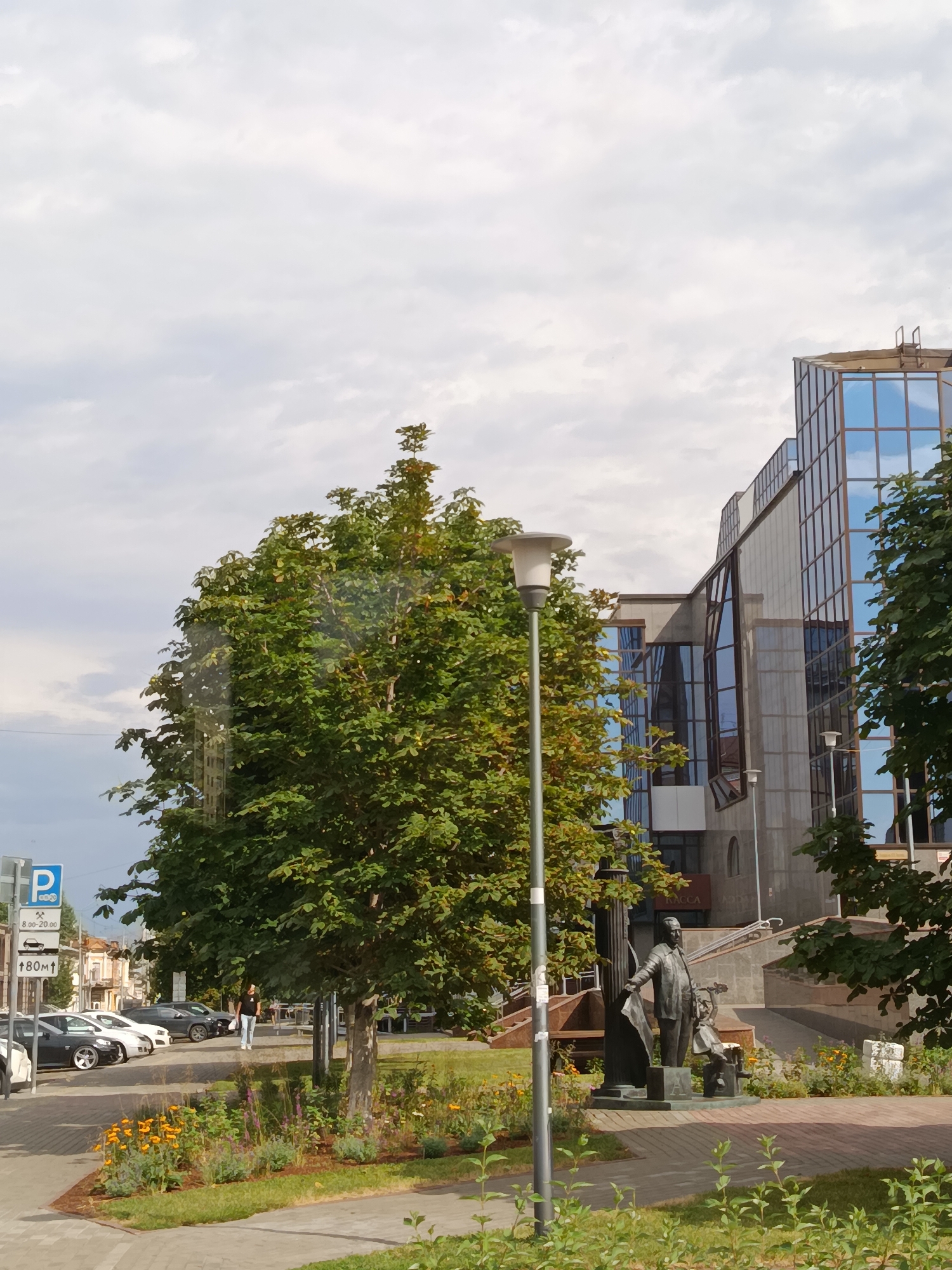 The image captures a street scene in Saratov, Russia, featuring a modern building with a glass facade, a lush green tree, and a statue. The building, with its blue-tinted windows reflecting the cloudy sky, suggests an urban or commercial area. A mature tree with dense foliage stands prominently in the foreground, partially obscuring a street lamp. To the right of the tree, a bronze statue depicts a standing figure holding a flag and a seated figure, situated on a stone pedestal. The statue is surrounded by a garden bed with blooming yellow and purple flowers. In the background, parked cars line the street, and a pedestrian crosses the sidewalk. A parking sign and a directional sign indicating "180m ↑" are visible on the left side of the image. The overall impression is a well-maintained public space with a blend of contemporary architecture and natural elements.
