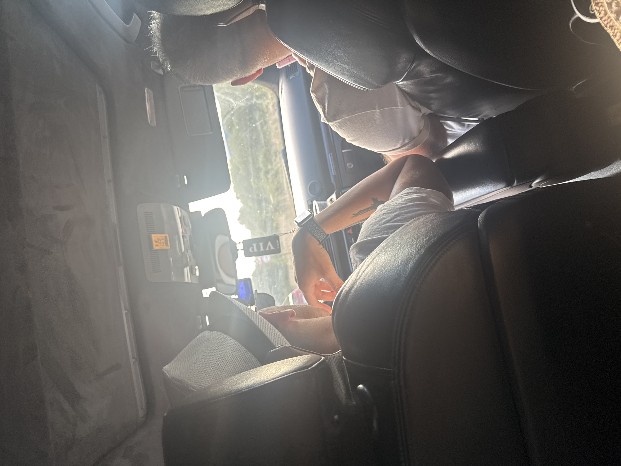 Two individuals are seated in the front seats of a car, facing forward. The person on the left wears a light-colored fedora and a white t-shirt. Their left arm is extended, and they appear to be gesturing. A watch is visible on their left wrist. A "VIP" tag hangs from the rearview mirror. The person on the right has light-colored hair and wears a light-colored t-shirt. They are looking down, possibly at a mobile device. The car's interior, including the dashboard and headliner, is visible. The setting appears to be a sunny day, with sunlight filtering into the car. The image is taken from behind the occupants. The scene is located in the city of Tsarevo, Bulgaria.