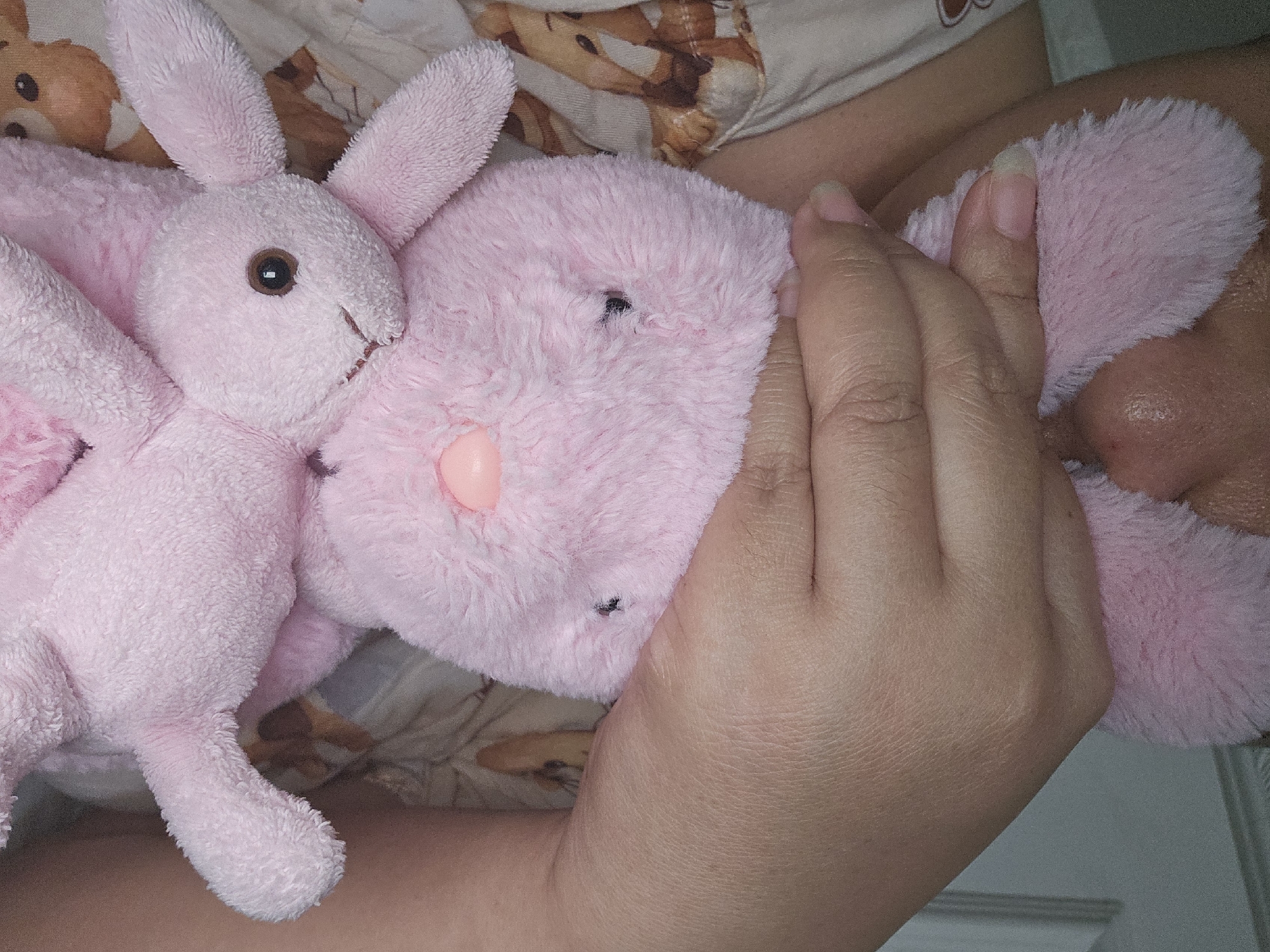A close-up shot shows a person's hand holding two pink plush bunnies. The larger bunny is in the foreground, with its head resting on the person's chest, and its ears are partially obscured by the hand. A smaller bunny is nestled beneath the larger one, with its head turned to the side. The person is wearing a light-colored shirt with a pattern of brown and white animal faces. The background is blurred, but appears to be a light-colored wall or door. There is no discernible location in Lamongan, Indonesia.