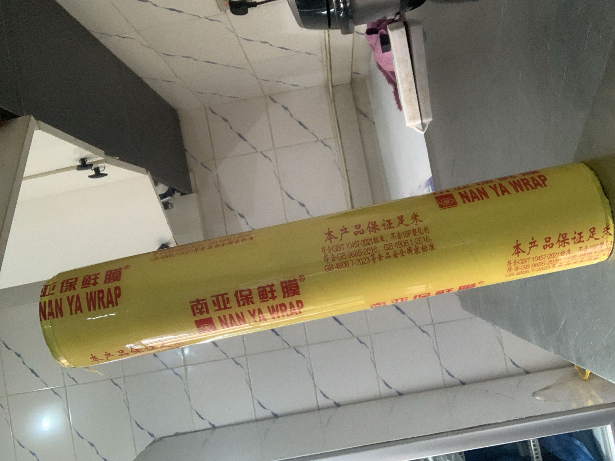 A roll of yellow plastic wrap with red Chinese and English text is leaning against a tiled wall in what appears to be a kitchen. The text on the wrap reads "NAN YA WRAP" in English and "南亚保鮮膜" in Chinese. Several lines of smaller text are also present. The environment suggests a domestic or commercial kitchen setting with white tiled walls and a dark countertop in the foreground. There are no people visible in the image. This scene is not located in Abuja, Nigeria.