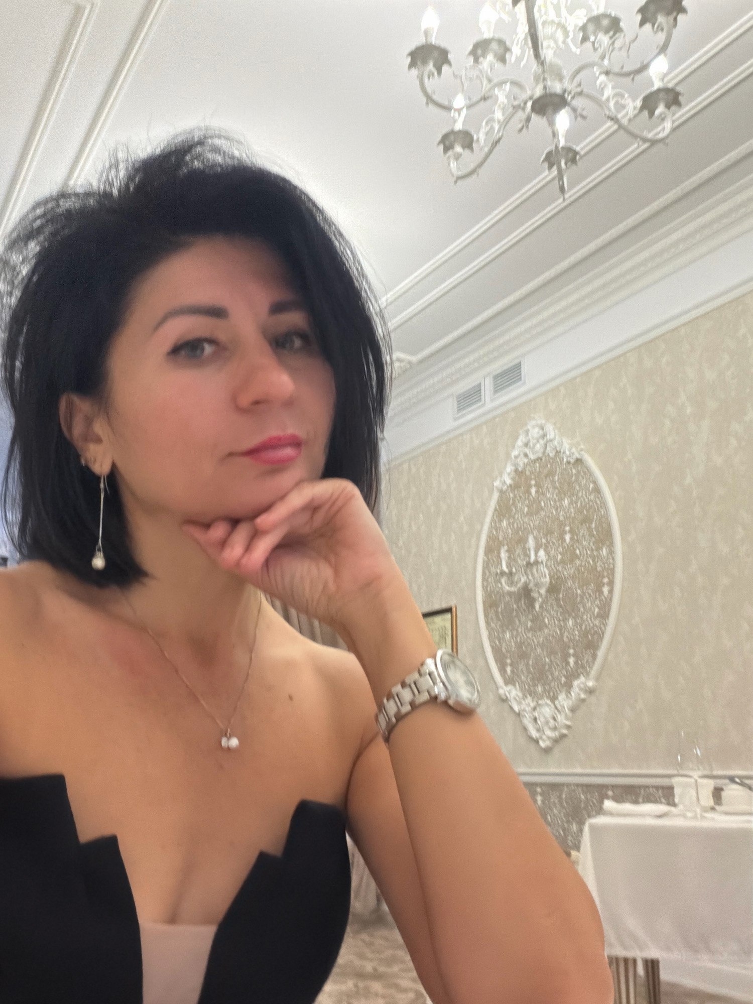 A woman with short, dark hair and medium skin tone is taking a selfie in what appears to be a restaurant. She is wearing a strapless black top with a tan underlay, a silver necklace with two pearls, and a silver dangling earring with a pearl. She is also wearing a silver watch on her left wrist. Her right hand is resting on her chin, and she is looking at the camera with her lips slightly parted. In the background, there is a chandelier, a decorative wall panel, and a table set for dining. The image does not contain any information that would allow for an identification of the location in the city of Angarsk, Russia.