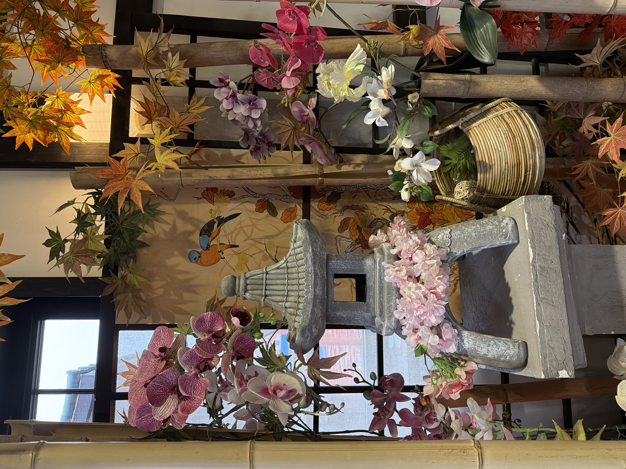 The image displays a densely decorated indoor scene, rich with artificial foliage and floral arrangements, evoking a Japanese aesthetic. Numerous faux maple leaves in vibrant orange, red, and green hues are interspersed throughout the display, creating an autumnal feel. These are complemented by a variety of artificial flowers, including bright pink and purple orchids, white and pale yellow blossoms, and clusters of delicate pink cherry blossoms, suggesting a mix of seasons.
Central to the arrangement is a prominent gray, stone-like replica of a traditional Japanese garden lantern (tōrō), adorned with pink flowers. Nearby, a spherical woven basket crafted from light-colored rattan or wicker holds additional green foliage and a decorative ball. Several natural bamboo poles are horizontally integrated, serving as structural or decorative elements that enhance the Japanese theme.
Behind these elements, a subtle mural or wallpaper features stylized tree branches and colorful birds, including a distinctive blue and orange bird. A window with a dark frame is visible on the left, allowing bright natural light to enter and revealing glimpses of a dark-tiled building roof outside. The overall composition is lush and layered, creating an immersive decorative environment.
The scene is a static decorative display, without any visible people or ongoing activity. No text is visible in the image. The location context provided for this scene is Khimki, Russia.