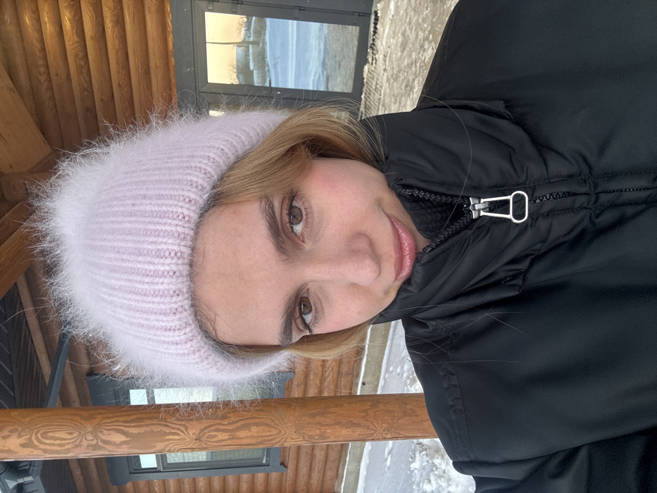 A female subject is depicted in a close-up, outdoor portrait under winter conditions. She has fair skin, light brown eyes, and light brown hair partially visible. She wears a light pink, fuzzy, ribbed knit beanie and a black puffy winter jacket with a high collar, partially zipped to reveal a silver zipper pull. Her expression is a subtle smile, and she gazes directly forward. The background features a log building to the left, constructed from light brown horizontal logs, with a dark-framed window and a vertical log support beam visible. To the right, a dark-framed glass door or window reflects a pale orange sky over a snow-covered landscape with indistinct distant features. Patches of snow and bare ground are visible beneath the subject. The location is an outdoor setting, indicative of a cold climate, near a log structure in Unknown, Unknown.