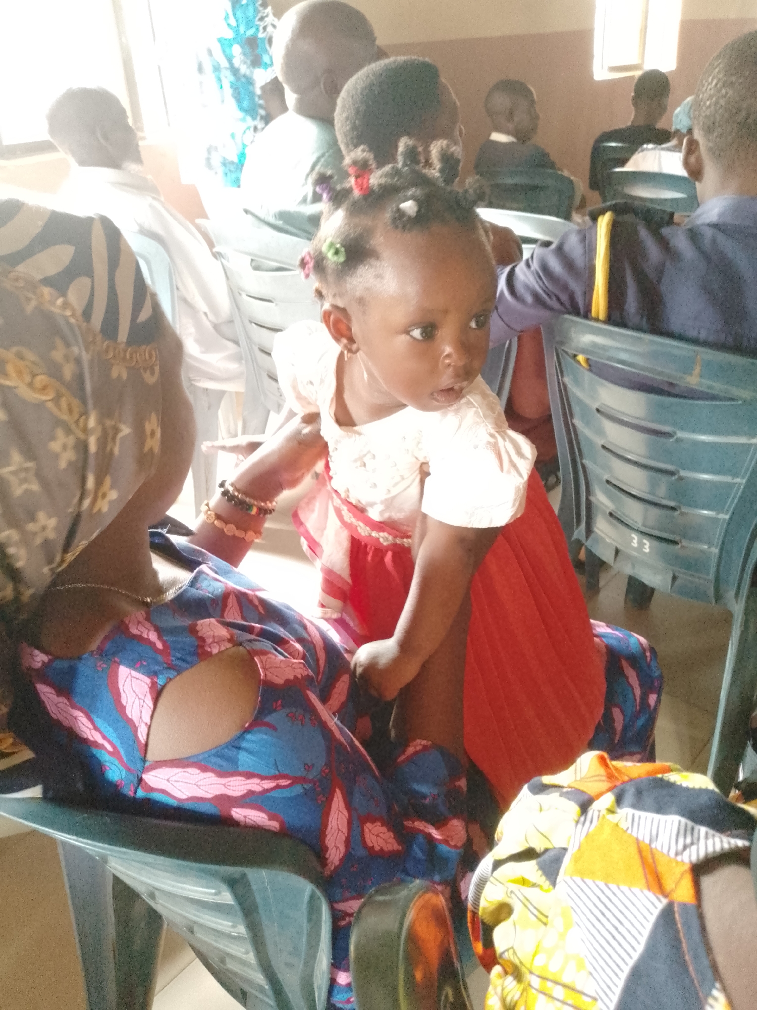 A medium shot shows a toddler with a red skirt and a white shirt standing on a woman's lap, the woman seated. The location appears to be indoors, with a group of people seated in chairs in the background. The toddler, who has small, decorated hair buns, is looking to the side. The woman has on a patterned blue and pink dress with a matching headwrap. There are other people in the background who are seated. All are in an unknown location.