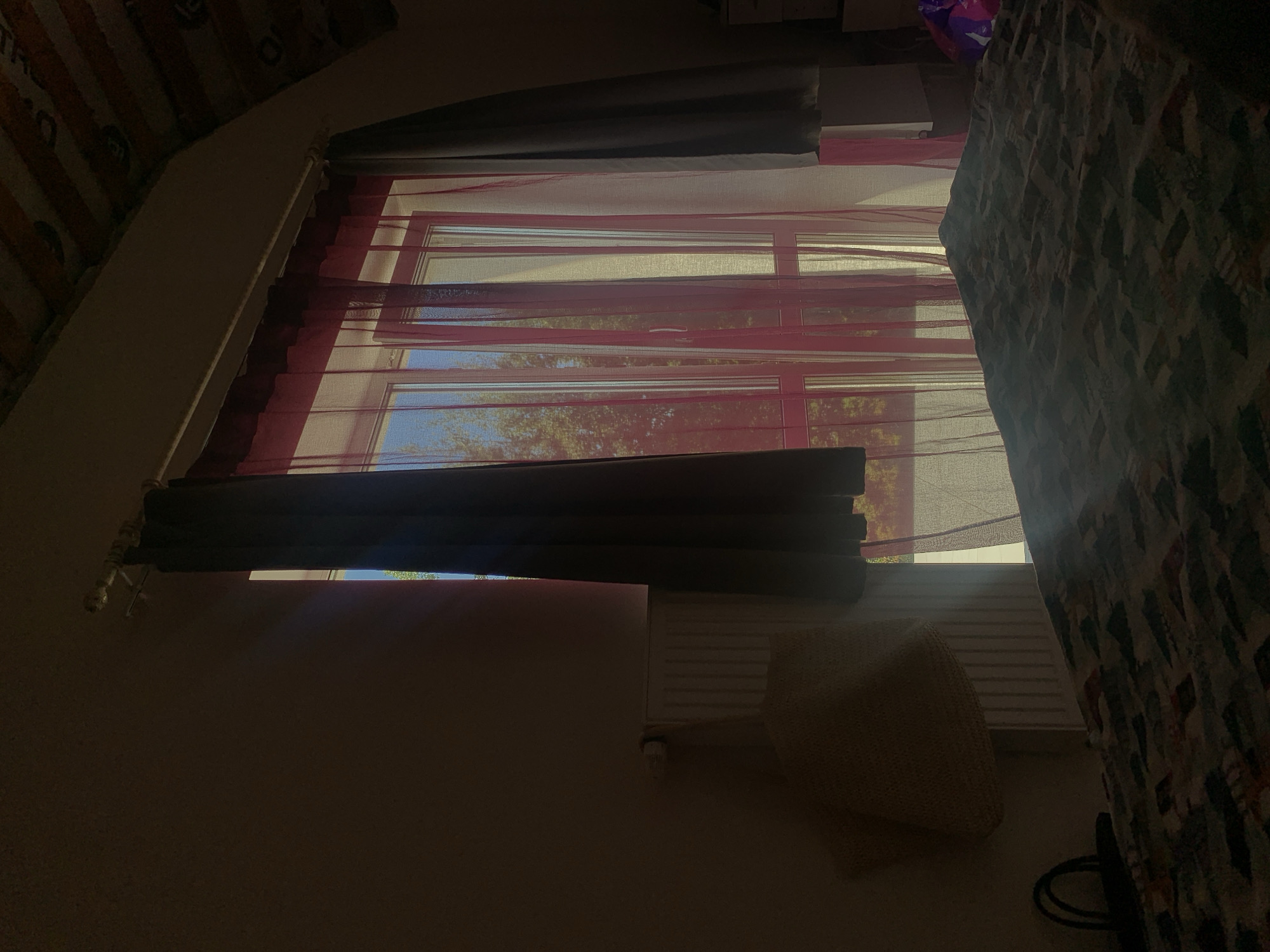 This is an indoor, eye-level shot of a bedroom with natural light streaming in from a window. The window is covered by a combination of dark grey curtains on the sides and a sheer pink curtain in the center, which diffuses the sunlight. The light casts a beam across the bed in the foreground, which is covered with a patterned duvet. To the left of the window, a white radiator is visible with a beige woven pillow hanging from it. The room has a slanted ceiling with exposed wooden beams. The overall impression is one of a quiet, domestic setting bathed in the warm glow of late afternoon sun. There are no people present in the image. The location is described as a bedroom in Minsk, Belarus.