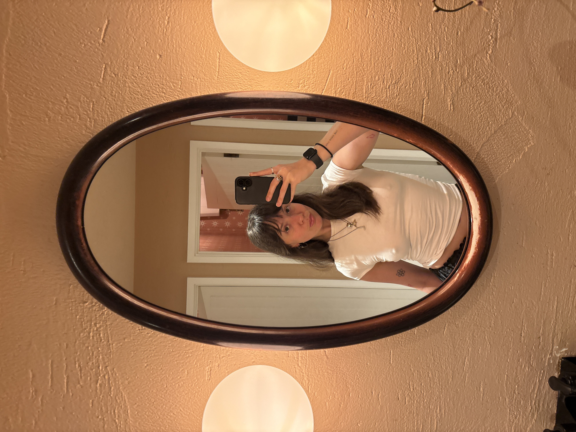 A young woman is taking a selfie in an oval mirror. She is holding a black smartphone with a triple lens camera in her right hand. She is wearing a white t-shirt and blue jeans. She has brown hair and bangs. She is wearing a watch on her left wrist and has tattoos on her left arm. The mirror is framed in dark brown wood and is mounted on a wall painted a peach color. There are two globe-shaped lights above and below the mirror. The reflection in the mirror shows a white door and a window with a red and white star pattern curtain. The location is unknown and could not be determined to be in London, United Kingdom.