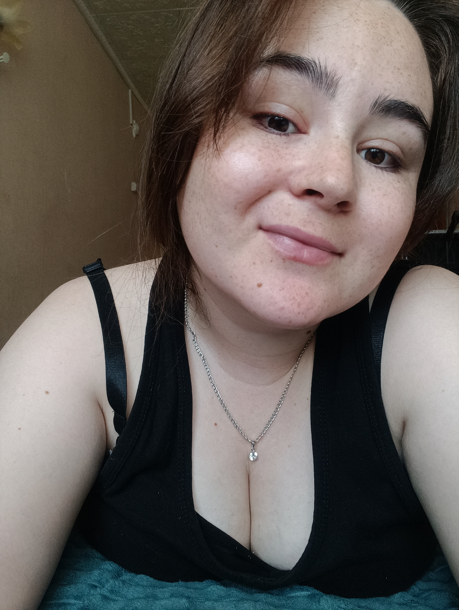 The image is a close-up selfie of a young woman. She has brown hair, brown eyes, and a light complexion with freckles. She is wearing a black tank top and a silver necklace with a pendant. The background appears to be an indoor setting with a plain, beige-colored wall. The woman is looking directly at the camera with a slight smile. There is no indication of her location in the city of Посад, Russia.