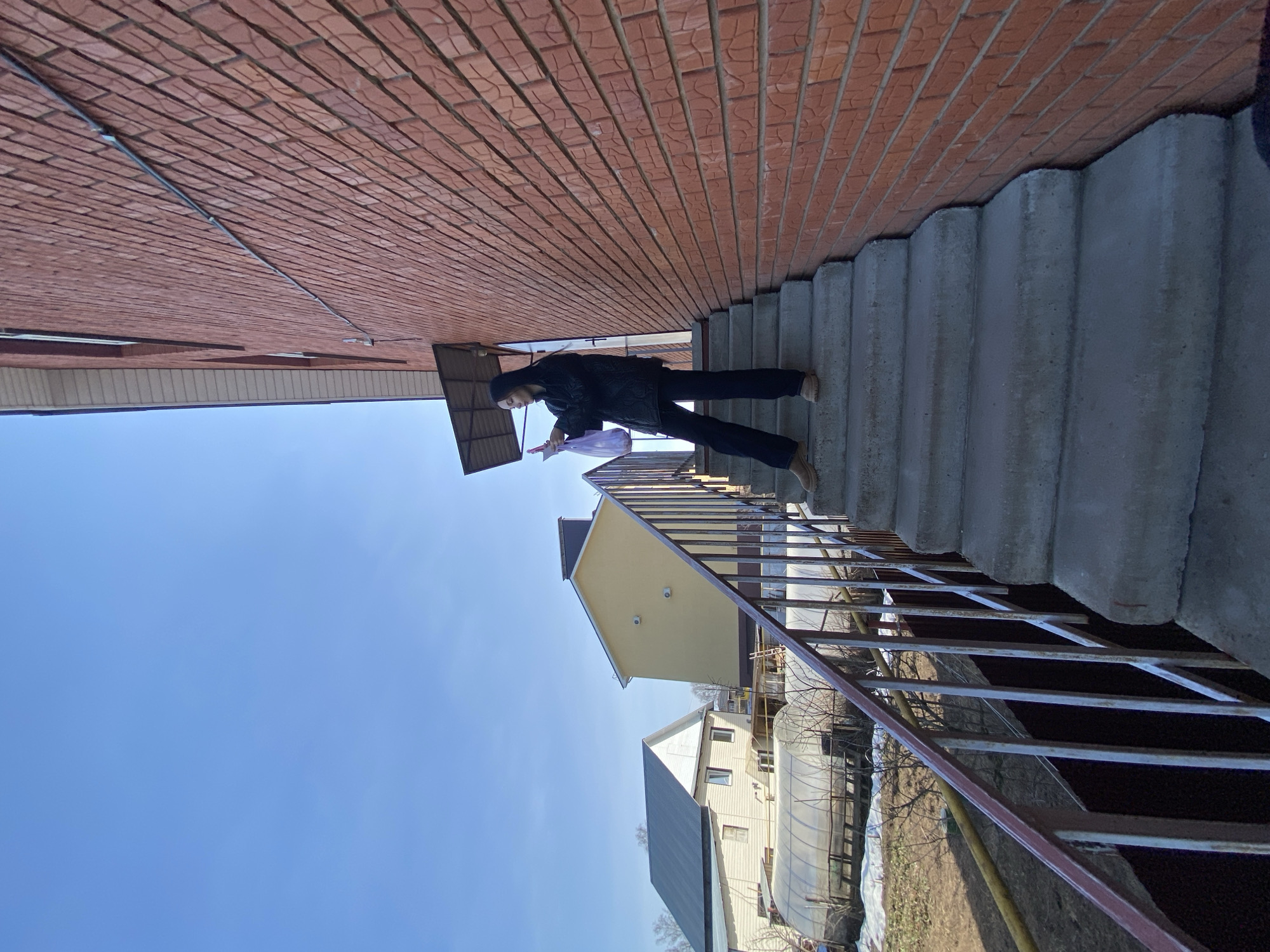A woman wearing a dark jacket, dark pants, and tan shoes is descending concrete stairs with a metal railing. She is holding a purple plastic bag. The stairs are adjacent to a brick building. In the background are houses and a greenhouse, under a clear blue sky. The location is likely Votkinsk, Russia, based on the request context.