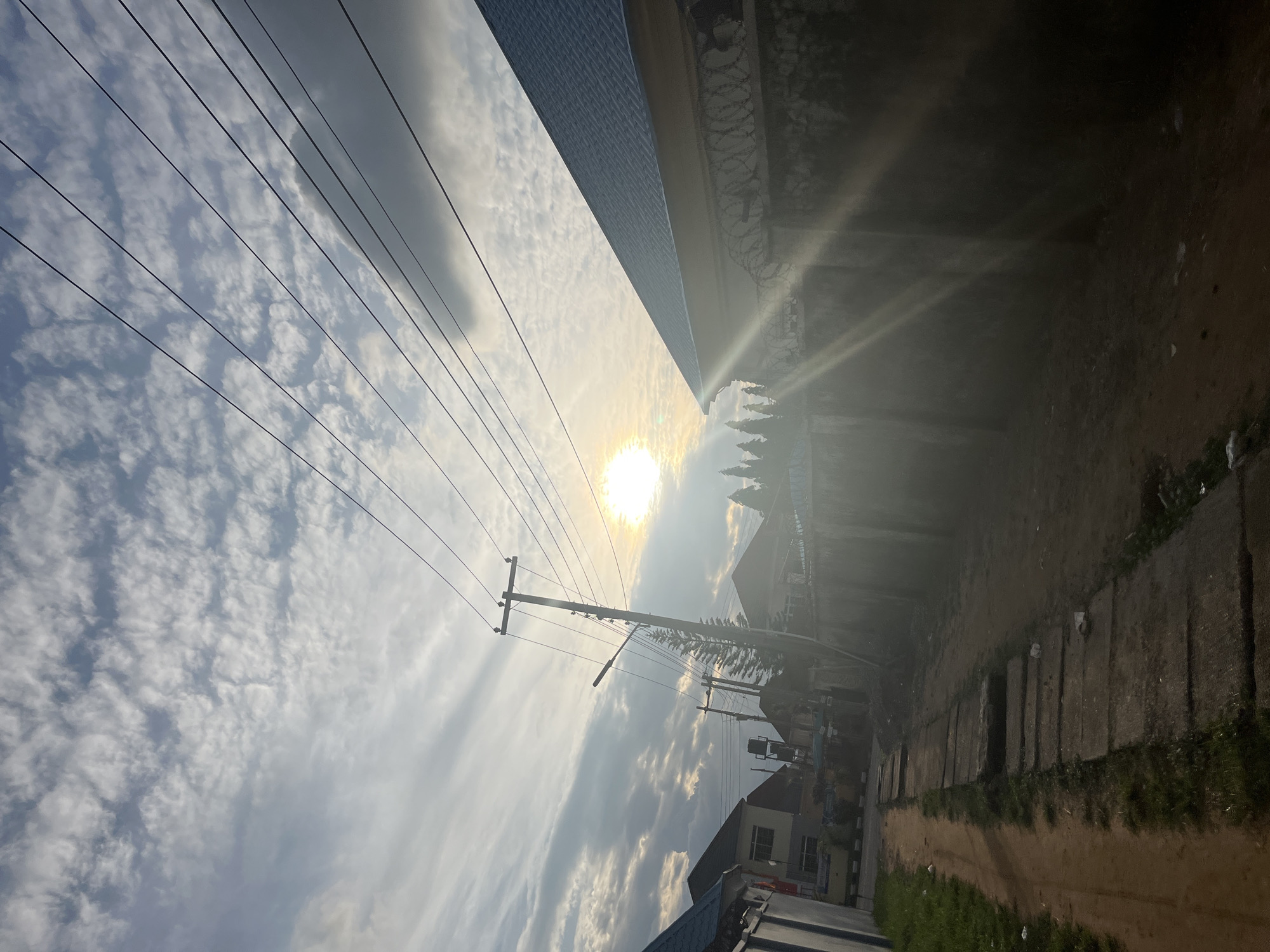 The image presents a sunlit, partially unpaved residential street scene in what appears to be a suburban area of Abuja, Nigeria. The sky is partially cloudy with scattered stratocumulus clouds. The sun is prominently visible in the upper-middle right quadrant, emitting strong rays that cause lens flare and backlighting across the scene. Multiple dark power lines extend diagonally across the upper sky, anchored to wooden utility poles. One primary utility pole stands tall in the middle-left, featuring a street light fixture, with a slender pine tree growing adjacent to it. Further in the background, another utility pole is visible.
On the left side of the frame, an unpaved dirt path runs from the foreground towards the background, bordered by sections of concrete slabs that function as drainage covers or an unfinished sidewalk, with patches of green grass interspersed. In the middle-left background, residential buildings with pitched roofs are partially visible. On the right side, a substantial building with a light blue/grey pitched roof dominates the upper-right portion. This building is fronted by a tall concrete wall, topped with a continuous line of coiled razor wire, which extends from the foreground into the mid-ground. The intense backlighting from the sun casts deep shadows across the lower right section of the image, obscuring details on the wall and ground behind it. A line of evergreen trees is visible behind the wall on the right. No people or animals are present in the scene. The lighting suggests either early morning or late afternoon.