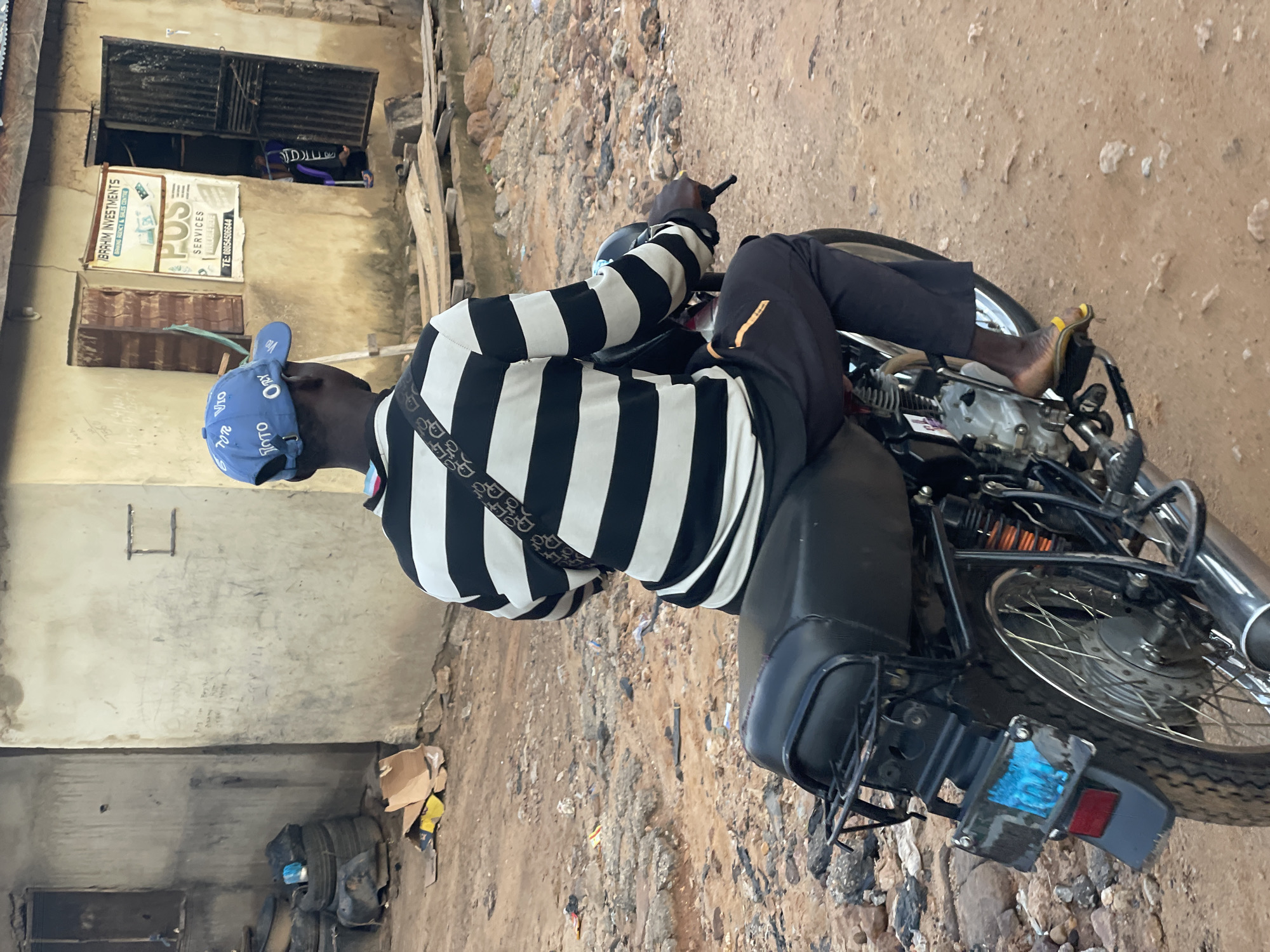 A man wearing a blue baseball cap and a black and white striped long-sleeved shirt sits on a black motorcycle, facing away from the camera. He wears dark pants and yellow sandals. The motorcycle has a black seat and a license plate covered in blue tape. The background shows a weathered, light-colored building with an open doorway, revealing a person inside wearing a purple garment. A sign above the doorway reads "IBRAHIM INVESTMENTS" and "POS SERVICES". The ground is dirt and gravel, with debris scattered around. The scene appears to be in a somewhat unkempt urban environment, possibly a street or alleyway in Lokoja, Nigeria, judging by the architecture and general setting.