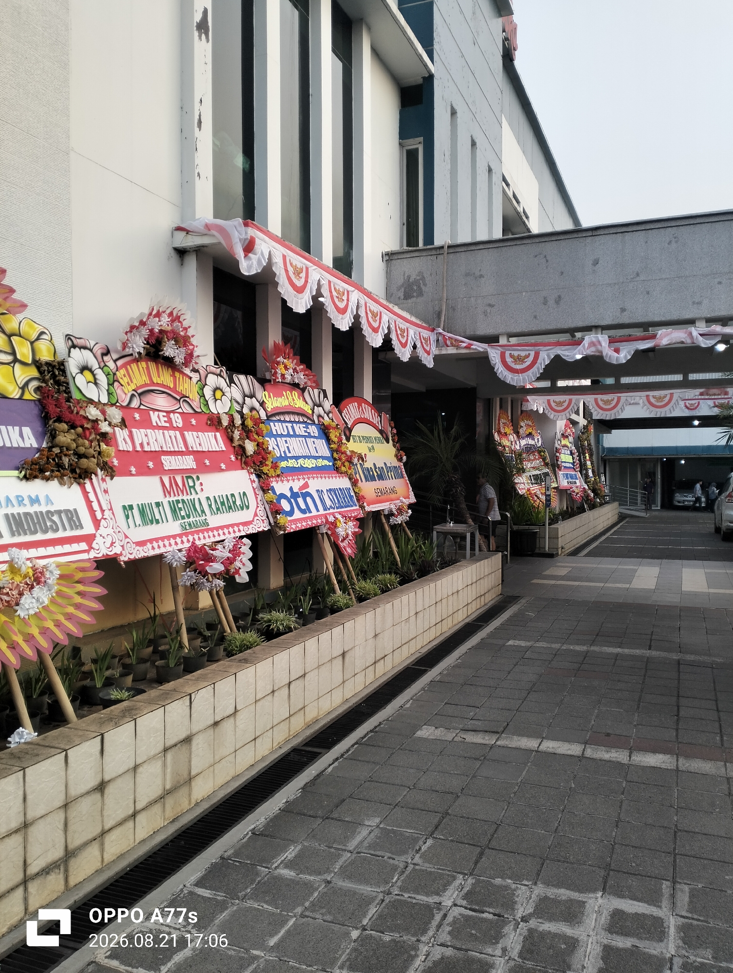 The image shows a row of floral arrangements, commonly known as "papan bunga" or flower boards, displayed outside a building with a white facade. These boards are decorated with colorful flowers and bear text, likely congratulatory messages or event announcements. The building appears to be a commercial or institutional establishment, with banners featuring Indonesian national symbols, such as the Garuda Pancasila emblem and red and white colors, adorning the entrance. The ground is paved with interlocking stone tiles, and a drainage channel runs along the side of the building. In the background, cars are visible in what seems to be a parking area or driveway, and a few individuals can be seen near an entrance. The overall scene suggests a formal event or celebration taking place at the location in Semarang, Indonesia.