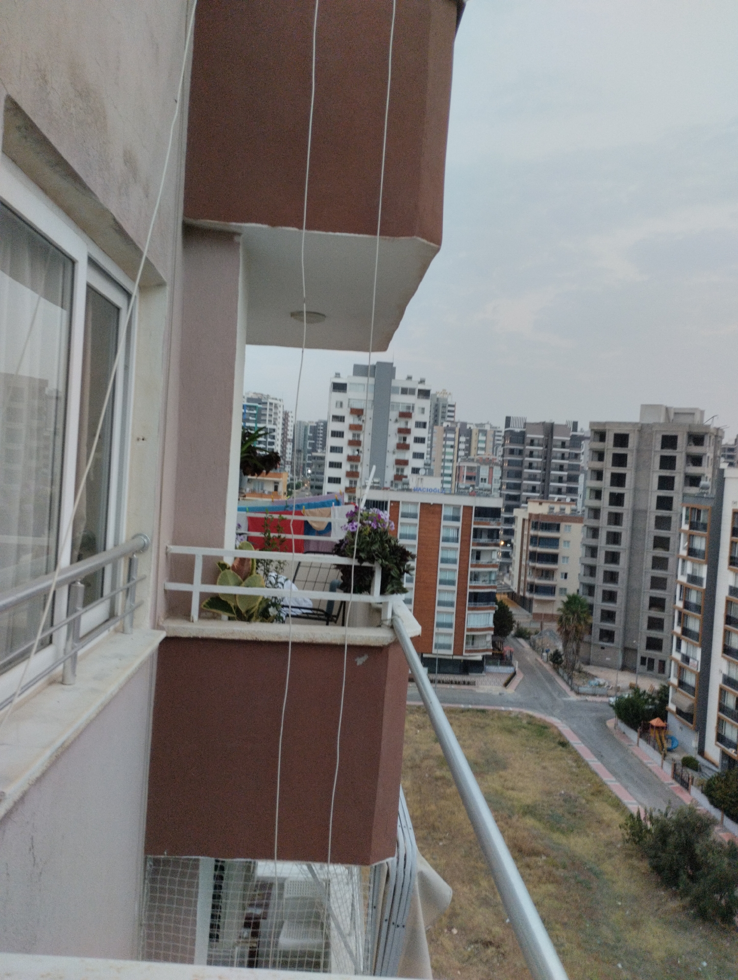 The image is a view from a balcony in Mezitli, Turkey, overlooking a city street and several apartment buildings. The balcony has a white railing and a brown exterior wall. On the balcony are potted plants, including a large green leafy plant and smaller flowering plants.
Beyond the balcony, the street is paved with a sidewalk. There are several tall apartment buildings of varying designs and construction stages. Some are completed and occupied, while others appear to be under construction with visible concrete frames and empty window openings. Greenery, including trees and grass, is visible in patches around the buildings and along the street. The sky is overcast with a light haze. A small playground area with a slide and climbing structure is visible in the distance. The overall impression is of a densely populated urban area with ongoing development.