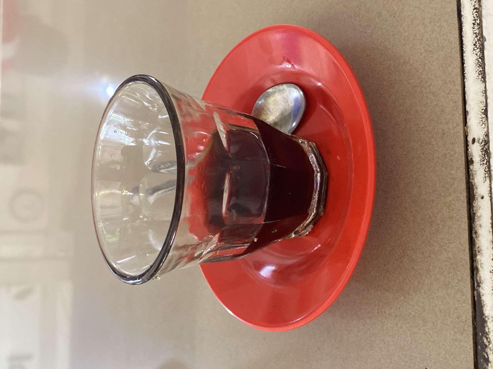 A close-up, eye-level shot shows a glass of dark liquid, likely coffee, sitting on a red saucer. The glass is made of clear, ridged glass and is about one-third full of the dark liquid. A silver spoon rests on the saucer to the right of the glass. The saucer is bright red and slightly concave. The entire setup sits on a textured, light brown surface that appears to be a table. In the blurred background, there are reflections and light patterns on a pale, vertical surface, suggesting an indoor setting. The scene is simple and focuses on the drink and its presentation. This image is taken in Lhokseumawe, Indonesia.