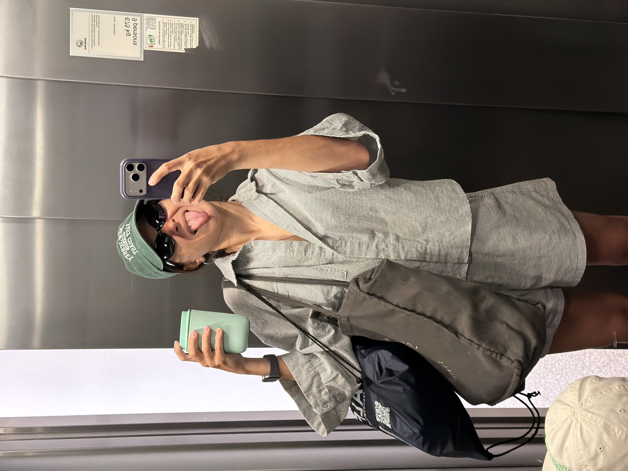 A woman stands in an elevator, taking a selfie with a phone in her right hand. She is wearing a green baseball cap, sunglasses, and a striped short set. Her tongue is sticking out. In her left hand, she holds a light green container. A gray tote bag and a dark blue drawstring bag hang on her shoulder. A document is affixed to the elevator wall, and the lower portion of another baseball cap is visible in the bottom right corner of the image. The elevator appears to be in Budva, Montenegro, based on the text present on the document affixed to the elevator wall.