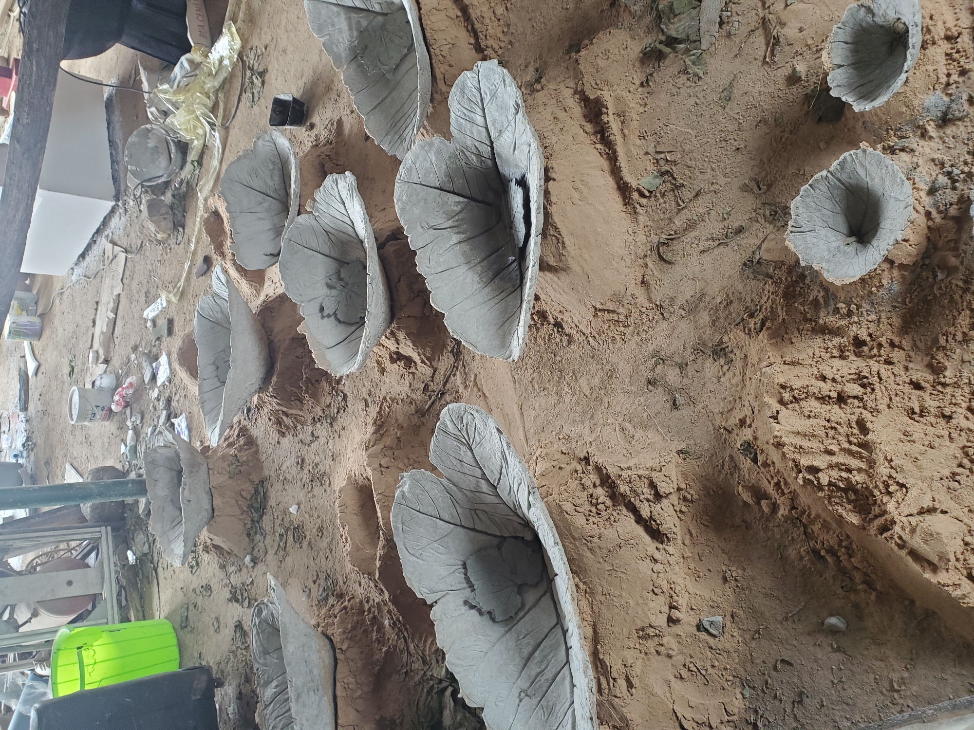 The image shows a collection of concrete leaf-shaped bowls resting on mounds of sand, likely outdoors in a workshop or construction area. The bowls are grey and detailed, resembling the veins of large leaves. They are arranged in a loose, scattered pattern on a dusty, sandy ground. In the background, there are various construction materials, including buckets, bags, and possibly furniture. The lighting suggests it is daytime. There are no people visible in the image. The scene depicts the unfinished stage of decorative items. The specific location in Jos, Nigeria, cannot be determined from the image alone, as it lacks any identifying landmarks or contextual clues specific to that city.