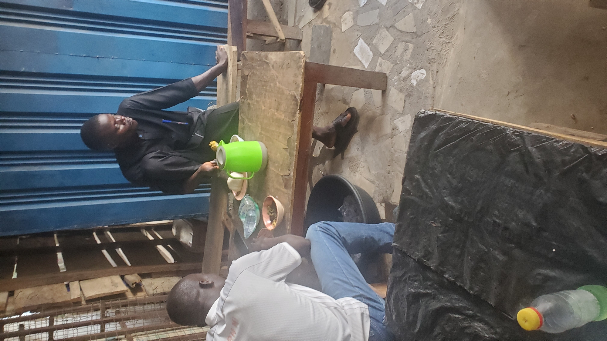 The image depicts an indoor scene, possibly a food stall or eatery, located in Jalingo, Nigeria. Two individuals are present. In the foreground, a person wearing a white polo shirt and blue jeans is seated facing away from the camera, appearing to be eating or preparing to eat from a large black basin. In the background, another individual, dressed in a black collared shirt, is seated at a wooden table. This table is laid with food items, including what appears to be fufu (a staple West African food), a green pitcher, and bowls of soup or stew. A blue corrugated metal sheet forms the backdrop. The floor is a mix of concrete and stone tiles. The overall setting suggests a casual, possibly rustic, environment.