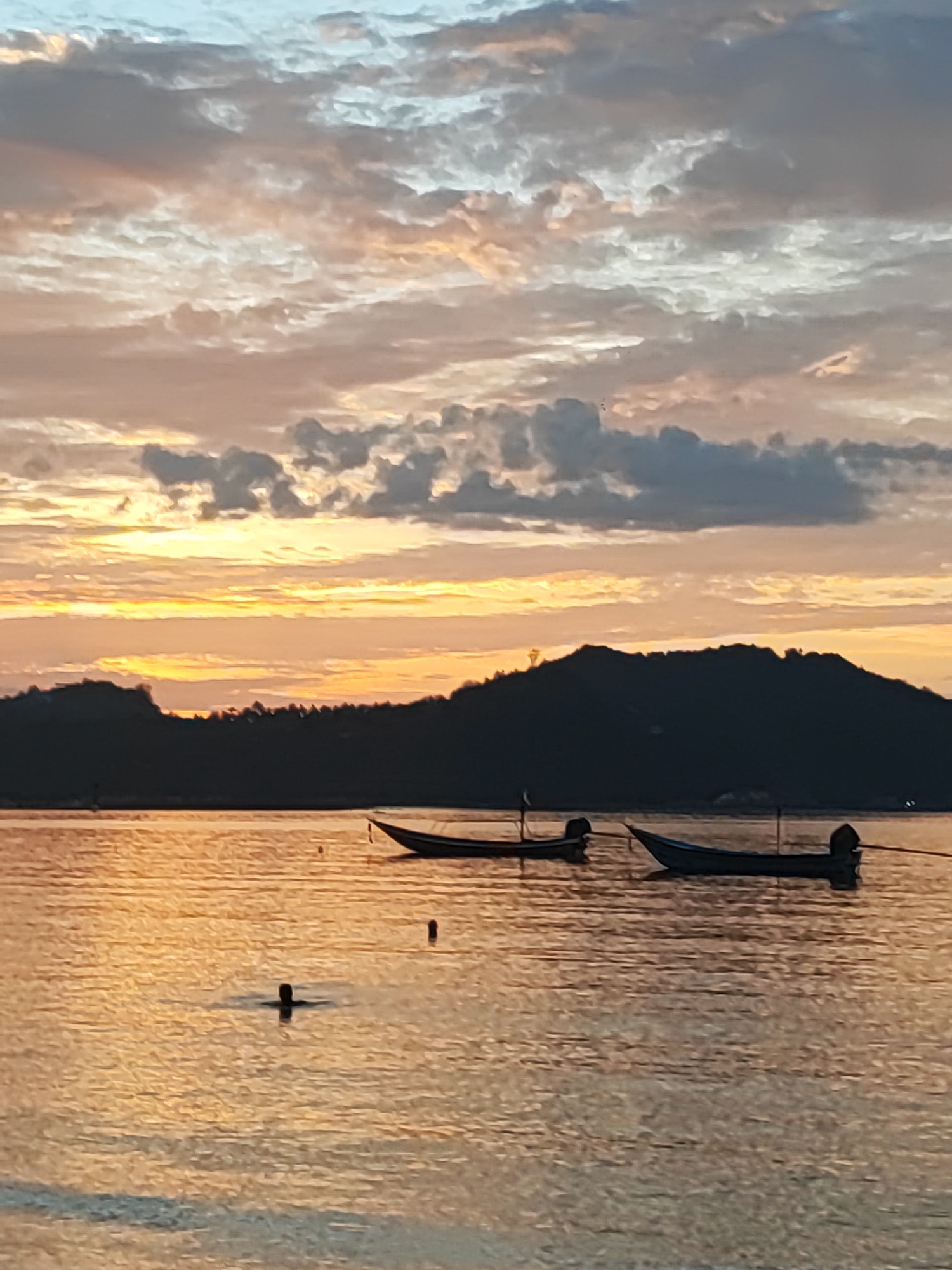 An outdoor, low-angle, vertical shot captures the serene scene of a tranquil bay in Ko Pha-ngan District, Thailand, during what appears to be a sunrise or sunset. The sky is a canvas of soft, muted colors, transitioning from pale blue at the top to warm oranges and yellows near the horizon, interspersed with scattered, wispy clouds. The silhouette of a lush, tree-covered hill forms a dark, undulating line against the vibrant sky.
In the foreground, the water reflects the sky's warm hues, creating a shimmering, golden surface. Two long, narrow boats are moored in the water. One boat, closer to the viewer and on the right, is a pale blue with a dark outboard motor. The other boat, to the left and slightly further away, is darker and also appears to have an outboard motor. A single person is visible in the water, with only their head and shoulders above the surface, appearing to be swimming or floating. The water is calm, with gentle ripples distorting the reflections. The overall impression is one of peace and natural beauty.