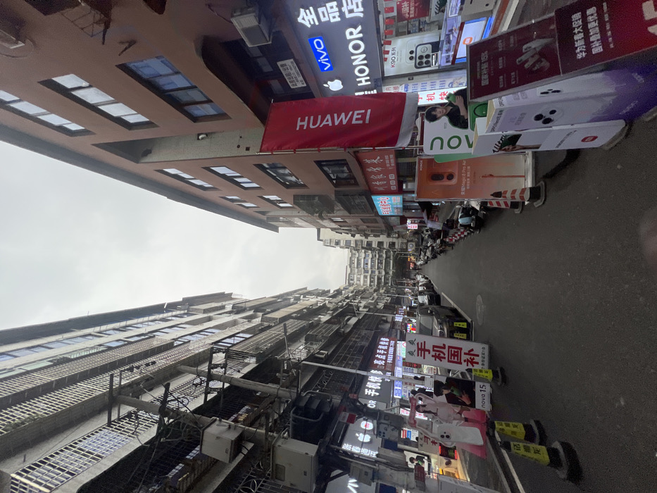 This image depicts a narrow urban street in Jiangjin, China, under an overcast sky. The perspective looks down the street, flanked by multi-story buildings on both sides. The building on the left is a taller, gray, concrete-faced structure with numerous windows, balconies secured with metal bars, and a dense network of overhead utility wires and external air conditioning units. The building on the right is a reddish-brown, multi-story structure with regularly spaced windows.
The street serves as a commercial thoroughfare, predominantly featuring mobile phone stores. Prominent signage includes a large red vertical banner displaying "HUAWEI" on the right building, a blue "vivo" sign, and a white "HONOR" sign. An Apple logo is visible on a shop front on the left. Numerous freestanding promotional display boards, primarily for mobile phone brands, are arranged along both sides of the street. These displays showcase images of various phones and models, with some text in Chinese characters, including "手机国补" (mobile phone national subsidy) and "nova 15."
The asphalt road surface appears damp. Several cars are parked along the left curb, while numerous electric scooters or small motorcycles are parked along both sides, particularly dense on the right. Yellow and black striped traffic cones are placed near promotional displays on the left, and red and white striped cones are visible on the right. No active people are clearly visible in the foreground or midground, although indistinct figures may be present within some shops. The overall scene suggests a busy commercial district.