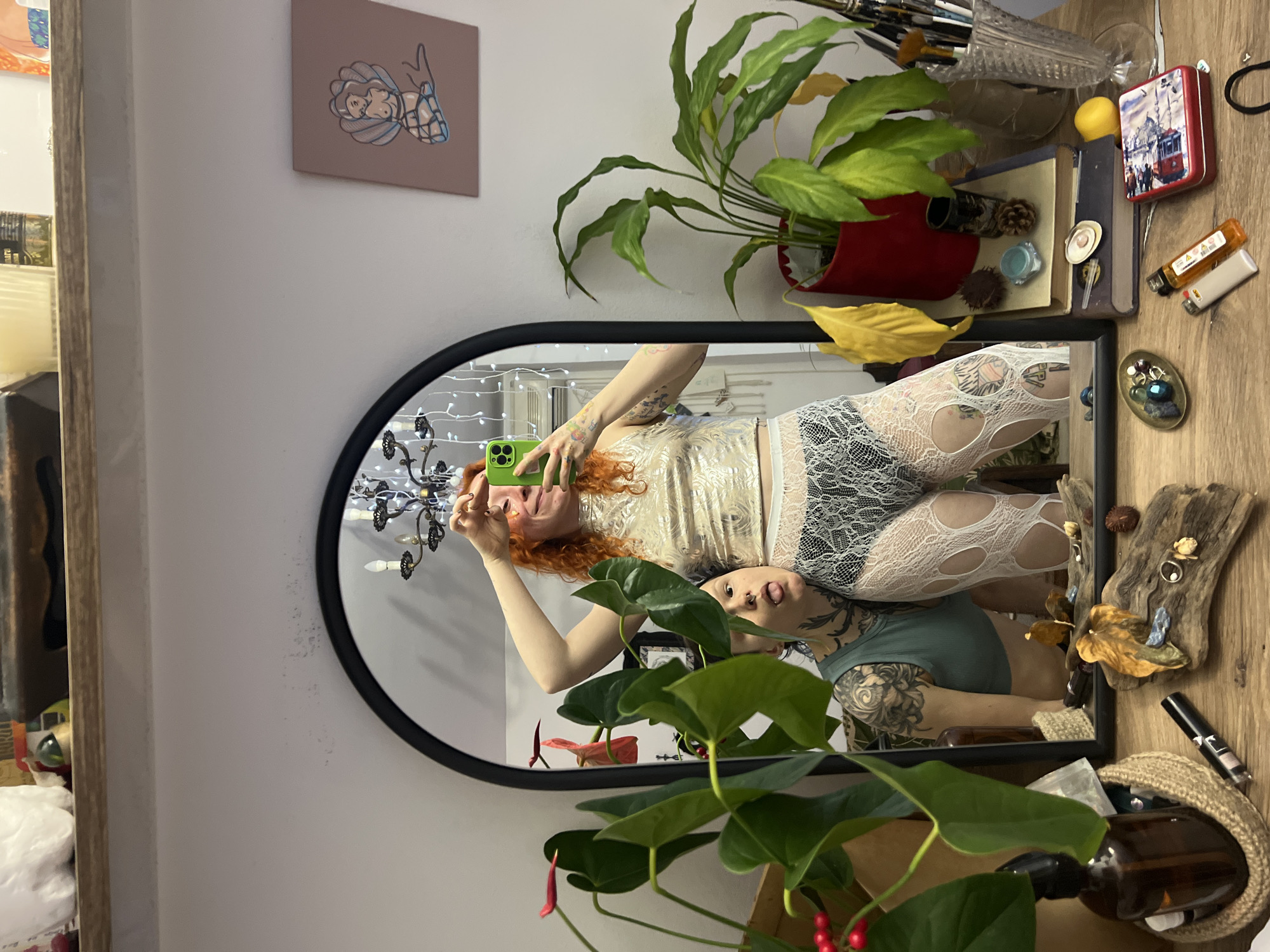 The image captures a mirror selfie. A person with red hair, wearing a sparkly top and lace leggings, holds a green smartphone, presumably taking the photo. The reflection in the mirror shows a second person wearing a green tank top with arm tattoos. Both appear to be inside a room. The mirror is surrounded by various objects, including plants, books, lighters, and decorative items. In the upper left corner of the image, there's a painting on the wall. Based on visual elements and the style, the image does not suggest a location within Belgrade, Serbia.