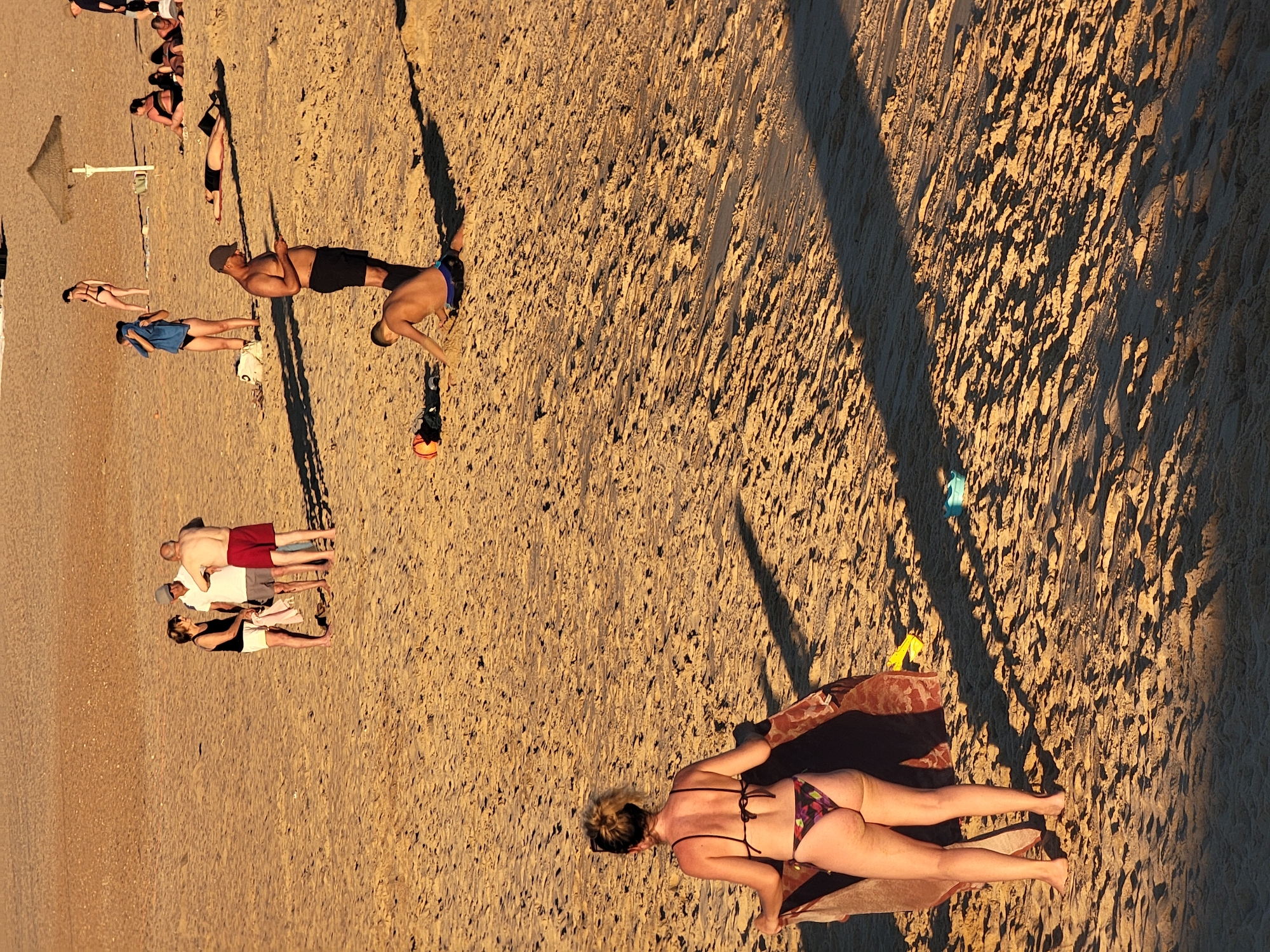 The image captures a sandy beach scene, likely in Quarteira, Portugal, with several individuals engaged in various beach activities. In the foreground, a woman in a patterned bikini stands facing away from the camera, holding a towel. To her right, a man wearing dark shorts and a baseball cap is looking down at a mobile phone, while a young man in blue swimming briefs kneels in the sand, appearing to play or build with it. Further in the background, a group of three people are conversing, with one man in red shorts and two others. Another person in a black bikini stands near a beach umbrella, and several other individuals are seen lying down or sitting on the sand, some partially obscured by shadows. The lighting suggests it is late afternoon or early evening, with long shadows cast across the sand. The environment is a typical beach with sand dunes and sparse vegetation visible in the distance.