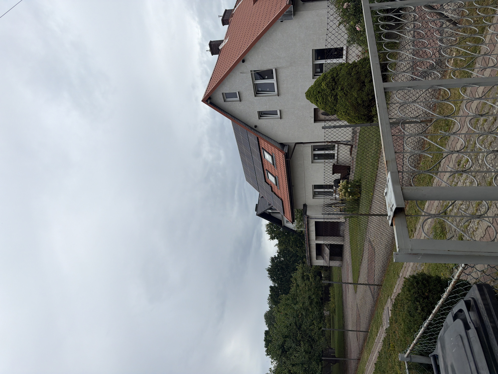 The image depicts a residential property located in Szczecin, Poland, under an overcast sky. The primary focus is on a light-colored two-story house with a red-tiled roof. The house features several windows, some of which appear to be newly installed with protective coverings. The roof of the house is partially covered with dark solar panels. Adjacent to the main house, a smaller structure, possibly a garage or outbuilding, is visible. A chain-link fence encloses the property, with a decorative metal gate in the foreground. The yard is landscaped with a well-maintained green bush in front of the house, and trees line the background. A black trash bin is situated near the fence on the left side of the image.