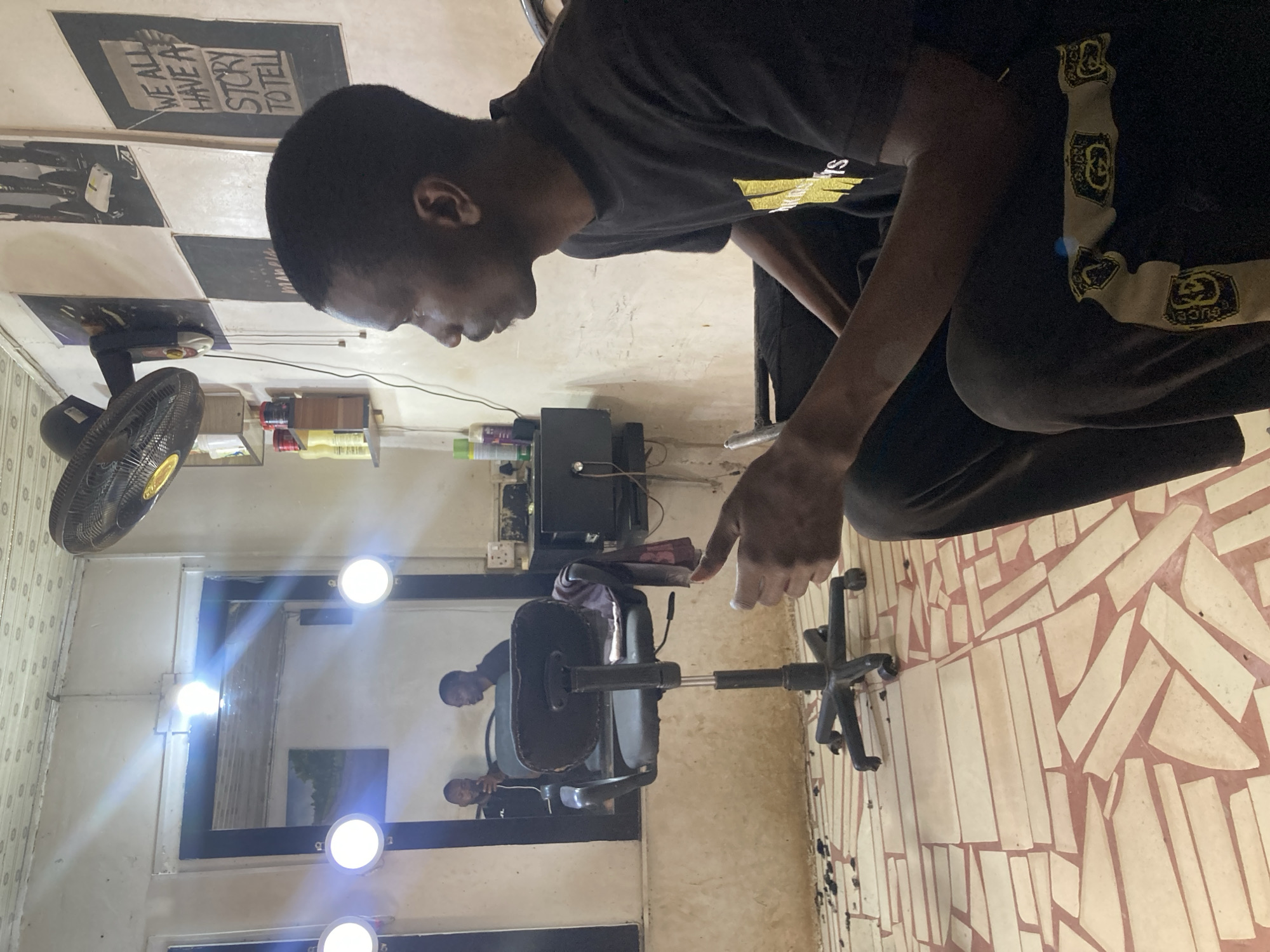 A man in a dark t-shirt and pants stands in a barbershop located in Gwong, Nigeria. The man is positioned near a swivel chair, his finger pointing towards the chair. The barbershop features a checkered ceiling, posters, a wall-mounted fan, and a large mirror reflecting the interior space with additional chairs and people. The floor is tiled with irregular patterns.