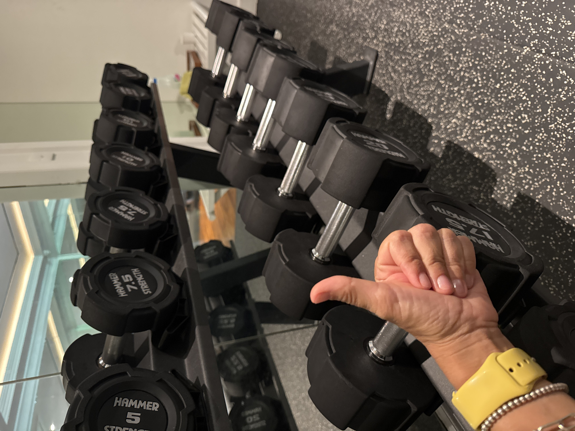 The image depicts a gym setting, showcasing a rack of dumbbells. The dumbbells are arranged in ascending order of weight. A hand with a yellow smartwatch is resting on one of the dumbbells. The background features a wall with a textured pattern and partial views of gym equipment. The precise location within Kuta Selatan, Indonesia, cannot be determined from the image alone.