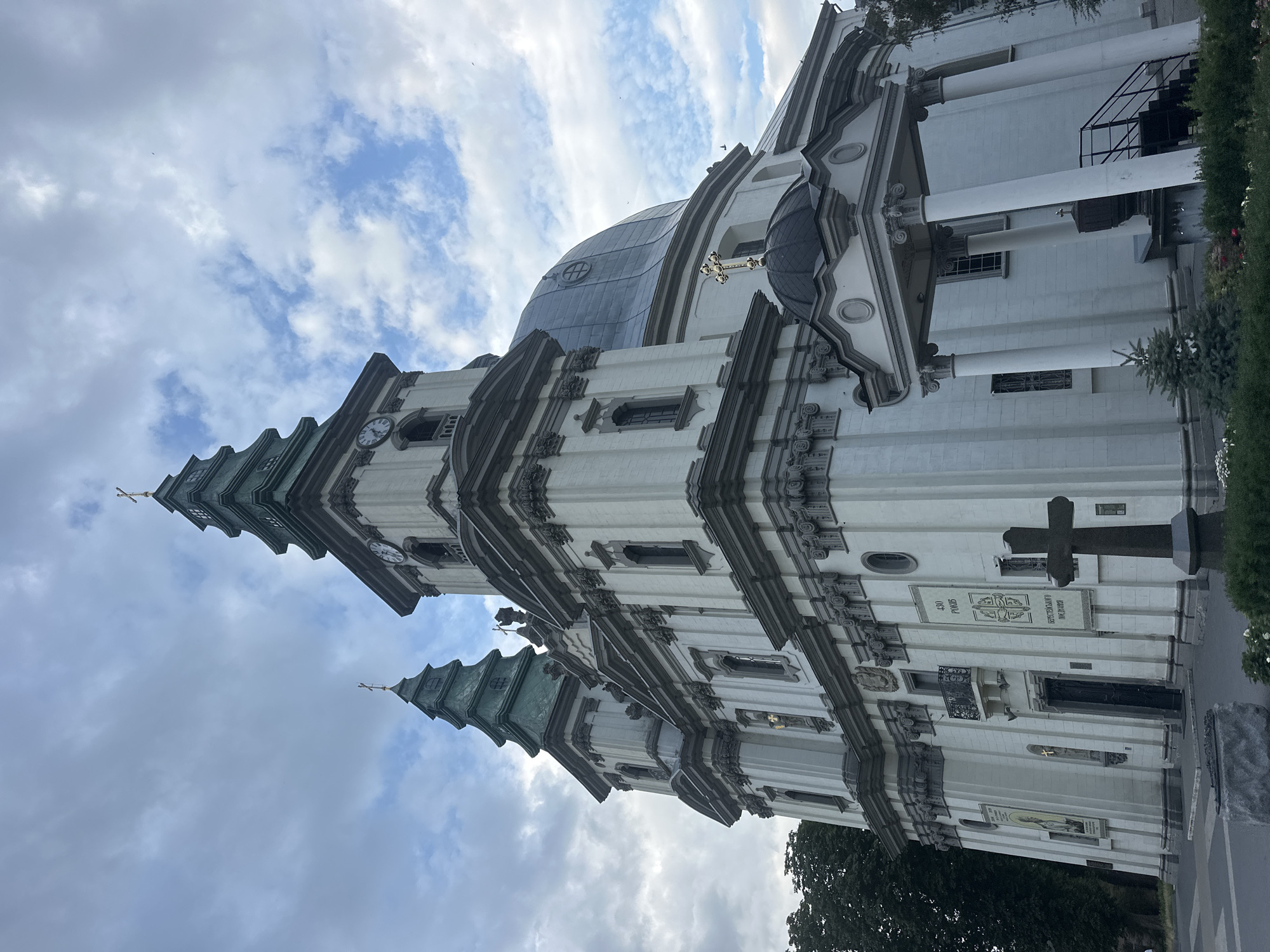 The image shows the Ternopil Cathedral, officially known as the Cathedral of the Immaculate Conception of the Blessed Virgin Mary, in Ternopil, Ukraine. The cathedral is a large, white building with a prominent dome and two tall towers topped with green spires. Both towers have clocks. A golden cross is visible on top of the dome. In front of the cathedral, there is a dark stone cross on a pedestal. The sky is partly cloudy with a mix of gray and blue. There are some trees visible to the left of the cathedral, and some bushes and small trees in the foreground. The perspective is from ground level, looking up at the cathedral. The image is taken during the daytime, under overcast conditions.