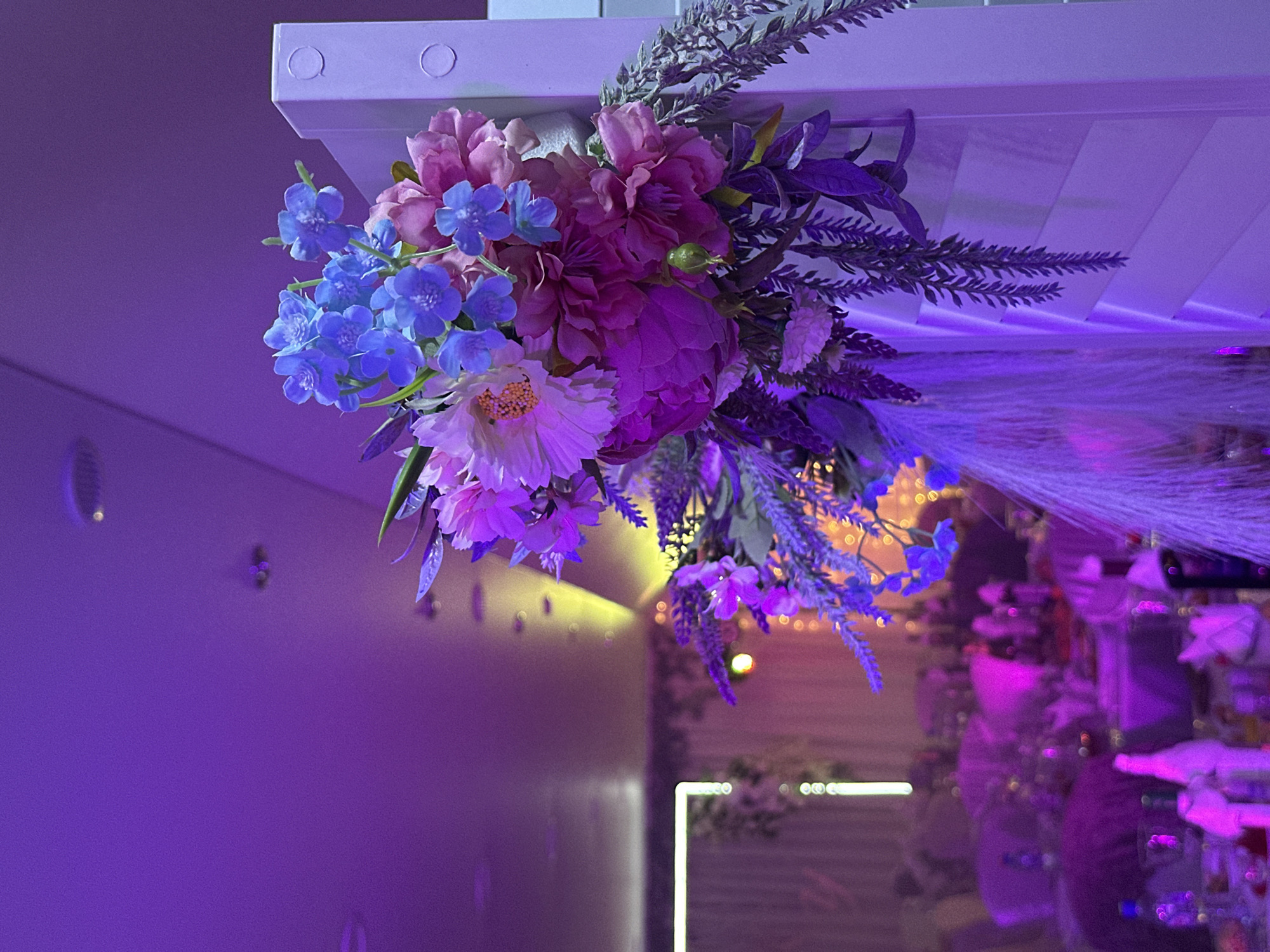 The image displays an indoor event venue in Chaikovsky, Russia, illuminated predominantly by purple and magenta ambient lighting, with a distinct yellow light beam cutting across the upper mid-ground. The foreground on the right features a vibrant artificial floral arrangement affixed to a white, vertical slatted panel, which also presents two small, circular indentations or features at its top right. The floral cluster comprises various artificial blooms, including distinct blue flowers resembling forget-me-nots, large magenta and pink peonies, a lighter pink flower with a visible yellow center, and spiky lavender-like elements. This is complemented by diverse artificial foliage, including dark green and purplish leaves and cascading fern-like fronds, alongside feathery, light-colored decorative plumes. The ceiling, visible in the upper frame, is plain and appears purple from the lighting, with several recessed spotlights. In the blurred background, multiple tables are set for an event, draped in light-colored tablecloths that reflect the purple illumination. Indistinct objects like bottles, glasses, and white napkins are visible on these tables. A glowing white rectangular light structure is present on the far left, and additional warm-toned bokeh lights are discernable deeper in the background. No people are visible within the scene.