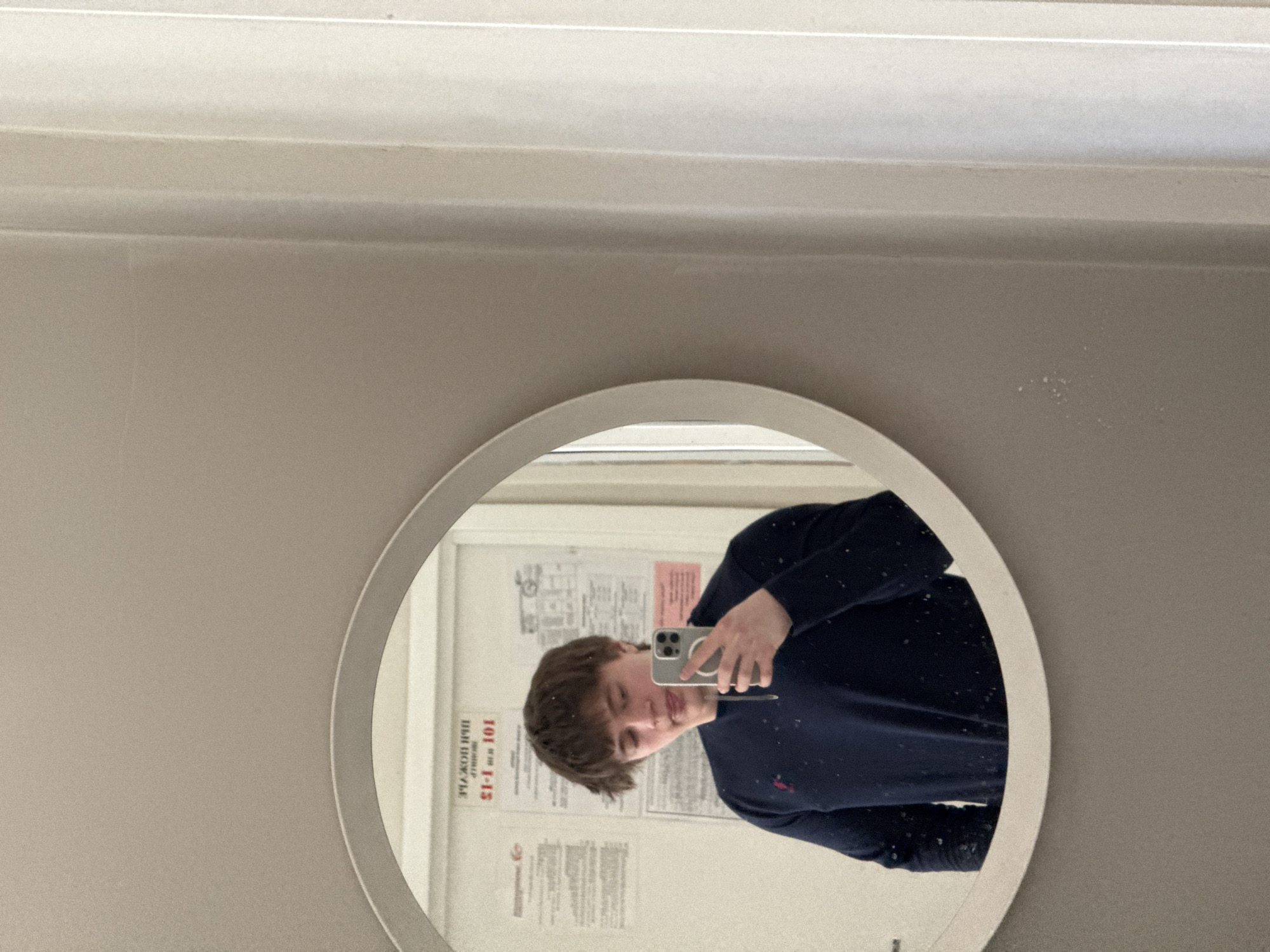 A person is taking a selfie in a round mirror. The mirror is mounted on a wall. The person is holding a smartphone. They are wearing a dark blue sweater. The reflection shows posters on the wall behind the person. Location is not discernible.