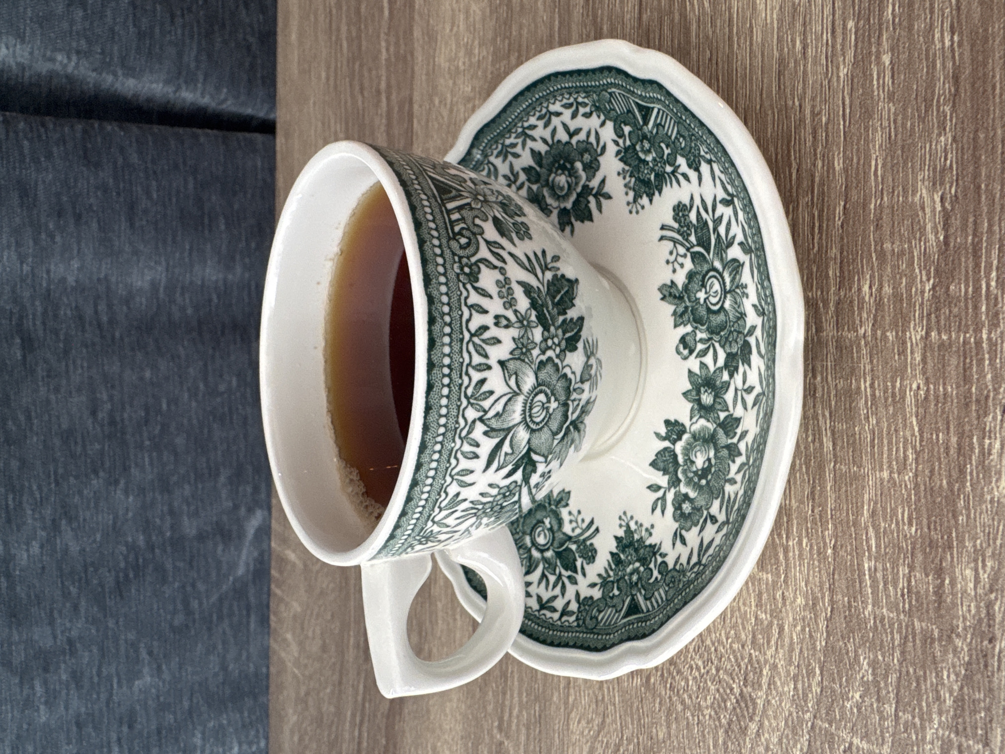 The image depicts a teacup and saucer set placed on a surface with a wood-like grain. The teacup is filled with a brown liquid, likely tea. Both the cup and saucer are white with a green floral pattern. To the left of the surface with the teacup and saucer, a darker blue-grey fabric or material is visible. The location is not discernible from the image. There is no indication of the scene being located in the city of Boreti, Montenegro. No people or actions are visible in the image.