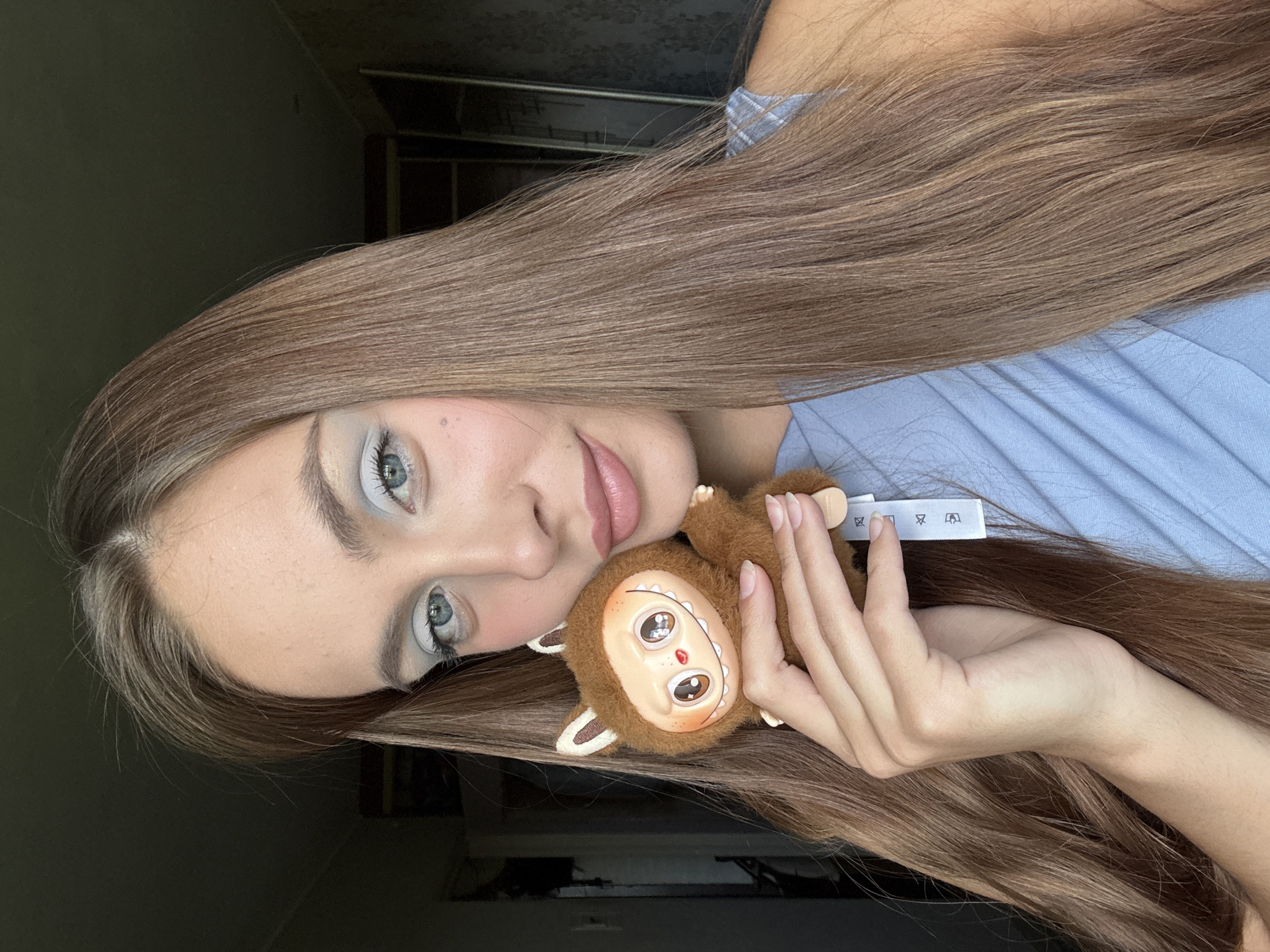 A young woman with long, light brown hair lies on her side, holding a small, fuzzy stuffed toy that resembles a monkey. Her hair cascades over her face, partially obscuring her features. She wears a light blue garment, possibly a shirt or dress, with a subtle plaid pattern on the collar.
The stuffed toy has large, round eyes with pupils, a small red nose, and a wide, friendly smile. Its fur is brown, and it has small ears. The woman's fingers are gently holding the toy. Her fingernails are long and appear to be painted in a neutral shade.
A white tag with black symbols is visible near the woman's hand, attached to her clothing. These symbols suggest laundry care instructions, with icons for washing, bleaching, ironing, and dry cleaning.
The lighting in the image is soft, suggesting an indoor setting. The background is dimly lit and appears to be a room with dark walls and possibly some furniture or shelving. The overall mood is relaxed and intimate, with the woman seeming to be comfortable and at ease. The time of day is not discernible, but the indoor setting and soft lighting do not suggest bright daylight. The image is a close-up, focusing on the woman and the toy, with the background out of focus. The location context provided is Шолохово, Russia, but there are no specific visual cues within the image to confirm this location.