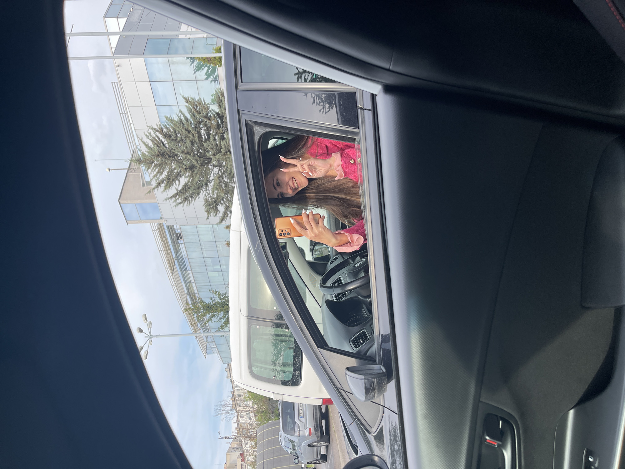 A bright, daytime image captures a joyful woman leaning out of the driver's side window of a black car, viewed from an adjacent vehicle. She smiles widely, her long brown hair flowing over her shoulders, and makes a peace sign with her right hand. In her left hand, she holds an orange smartphone, possibly taking a picture or video. She is wearing a vibrant pink top with ruffled accents.
The background reveals a clear blue sky with some white clouds and modern buildings featuring large glass windows, suggesting an urban or business district. A green coniferous tree stands prominently in the middle ground, adding a natural element to the scene. Several vehicles are parked nearby, including a white van and a gray SUV, the latter displaying a partially visible Bulgarian license plate "C 5438 BP," confirming the location in Sofia, Bulgaria. The foreground shows the dark interior of the car from which the photo is taken, including parts of the door frame and interior trim. The overall mood is cheerful and friendly, depicting a casual interaction.