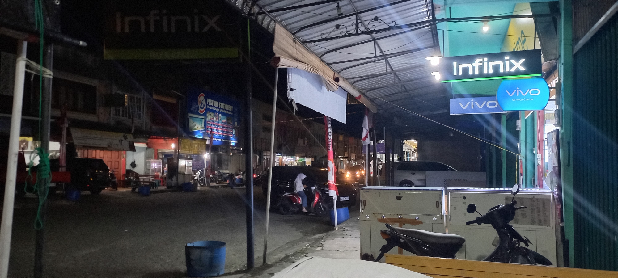 The image depicts a street scene at night in Tapak Tuan, Indonesia, possibly a commercial area with several shops and signage. Dominant elements include illuminated signs for "Infinix" and "vivo Service Center," indicating the presence of mobile phone retailers or service providers. A "Pestone Stationery" sign is also visible, suggesting a stationery shop.
The environment is characterized by a covered walkway or awning over the storefronts, illuminated by overhead lights. The street in front is paved and appears to be relatively quiet, with a few parked motorcycles and a black car. One motorcycle has a person, a woman wearing a white hijab, sitting on it. Another person, also wearing a hijab, is visible on a separate motorcycle in the foreground.
Objects present include blue trash bins, wooden tables and chairs outside a shop, and what appear to be display cabinets or refrigerators. The overall lighting suggests dusk or nighttime conditions, with artificial lights providing the primary illumination. The architecture of the buildings seems to be typical of Indonesian urban commercial streets.