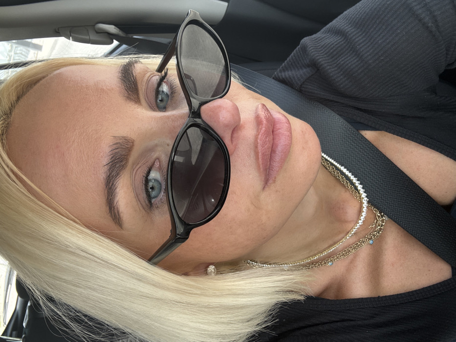 A close-up image depicts an adult individual, likely female, seated inside a vehicle. The subject has short, straight blonde hair and blue eyes, partially visible above black-framed, dark-tinted cat-eye sunglasses. Dark, well-defined eyebrows are present, and the lips appear full with a glossy finish. A small, clear stud earring is visible on the left earlobe. The subject is wearing a black, ribbed, long-sleeved top and a black seatbelt diagonally across the chest and shoulder. Multiple layered necklaces adorn the neck, including a choker-style necklace with small white stones and several gold-toned chain necklaces, one featuring small blue spherical beads. The subject is looking directly towards the camera. The background, seen through a vehicle window, is blurred, showing hints of light-colored structures, suggesting an outdoor urban or suburban setting under bright conditions. No explicit visual cues within the image confirm the location as Dubai, United Arab Emirates.