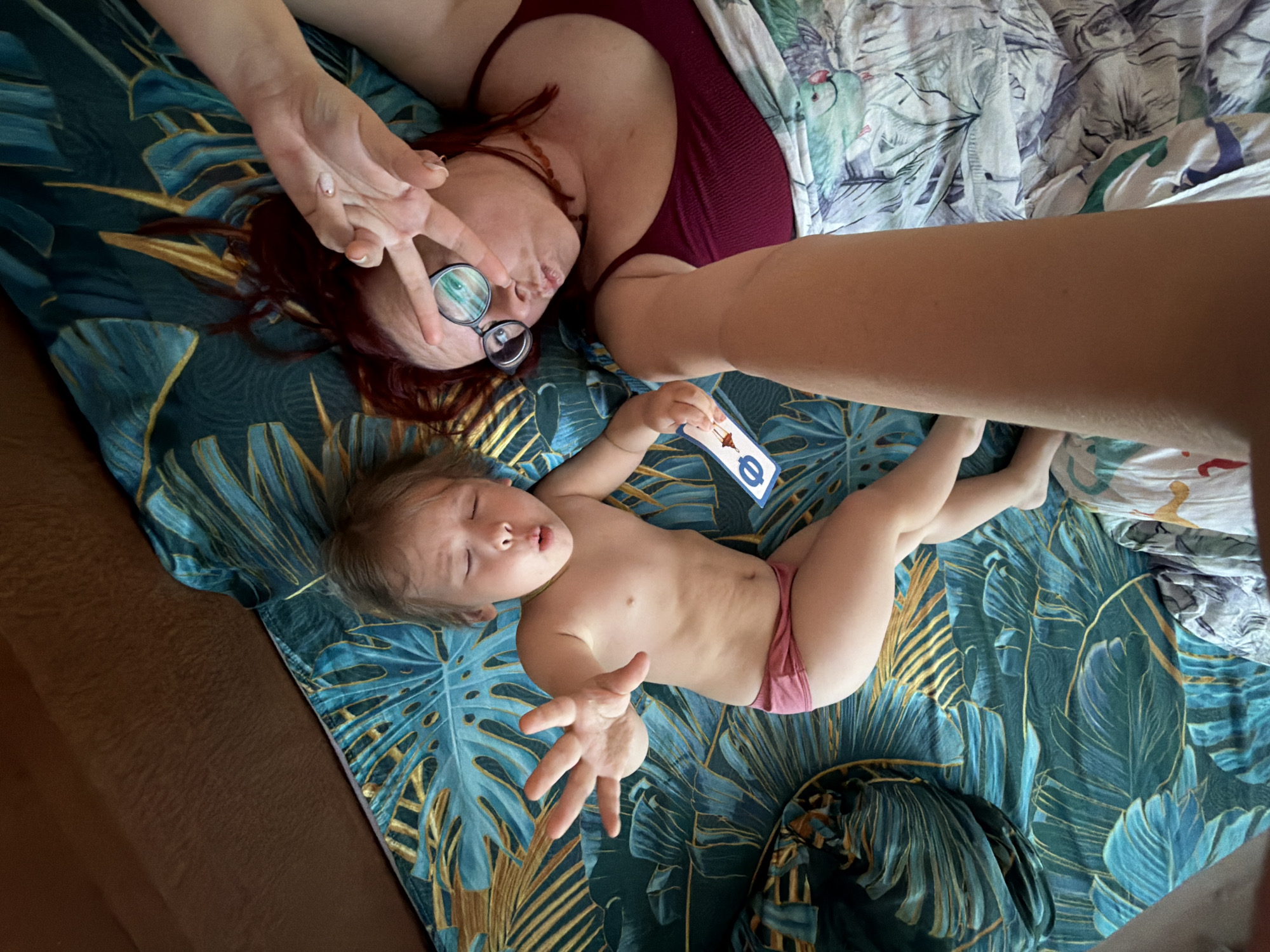 A top-down shot depicts a woman and a young child lying on a bed with blue and gold palm-print bedding. The woman, wearing glasses and a maroon top, makes a peace sign. The child, wearing pink underwear, holds a card with a London Underground logo. A draped cloth with a similar palm print design and another bed with a different print are partially visible. The location, Chaikovsky, Russia, cannot be verified from this image alone.