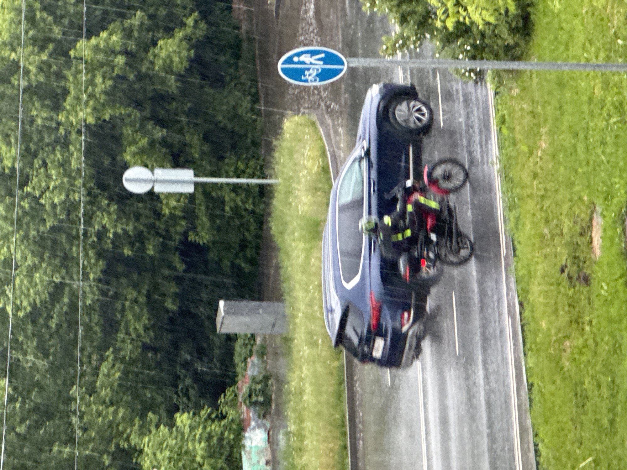 This media file presents an overhead, rotated view of a road scene in Perm, Russia, depicting what appears to be a traffic incident. A dark blue SUV, possibly a BMW, is stationary on the wet asphalt road, near a curb lined with grass and trees. Its front passenger wheel is visibly turned.
Adjacent to the SUV's rear passenger side, a black and red motorcycle or scooter is seen, suggesting it may have fallen or is being secured. Two individuals, clad in black uniforms with prominent yellow/green reflective stripes characteristic of emergency services or road assistance personnel, are attending to the motorcycle; one person is actively leaning over it.
The road is a two-lane asphalt surface with white lane markings, visibly wet from rain or recent precipitation. Lush green trees and grass line both sides of the road. Above the road, a blue circular sign with white pictograms indicates a shared path for pedestrians and cyclists. Another white, rectangular informational sign is visible on a separate pole. Several utility lines run across the upper left portion of the image.
The incident occurred during daylight hours, under rainy or recently rainy conditions. The car's rear license plate is partially visible, showing blurry characters including what appears to be "P" and possibly "5" or "S". No other discernible text is visible on signs or uniforms.