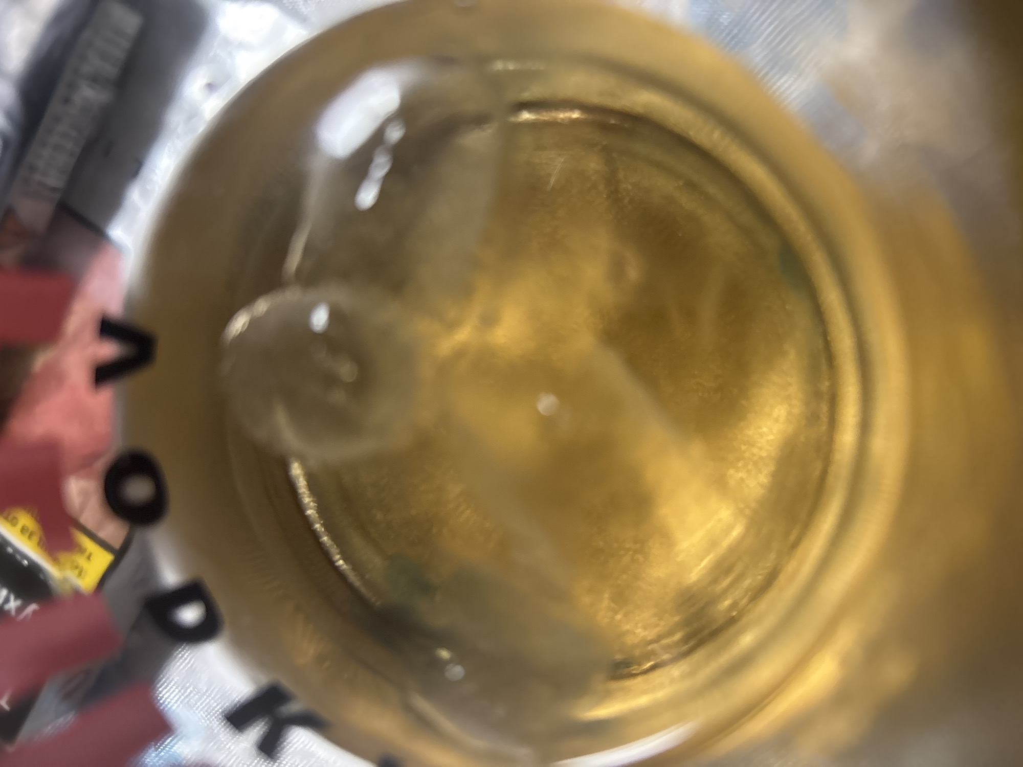 The image is a close-up, overhead view of a beverage in a glass. The liquid is a golden-brown color, and there are visible solid particles and cloudy wisps within it, suggesting it might be unfiltered beer or another sediment-containing drink. There appear to be ice cubes or frozen elements submerged in the liquid, with some distortion of light and reflection around them. The glass is transparent, and the outer surface shows some texture and reflections. In the blurred background, elements of packaging are visible, including text that appears to be in Cyrillic script and part of a label with yellow and black elements. One discernible letter from the background text is "V". The overall impression is that of a drink being consumed in an informal setting, possibly outdoors or in a bar. There is no discernible information in the image that indicates a specific location within Trudovets, Bulgaria.