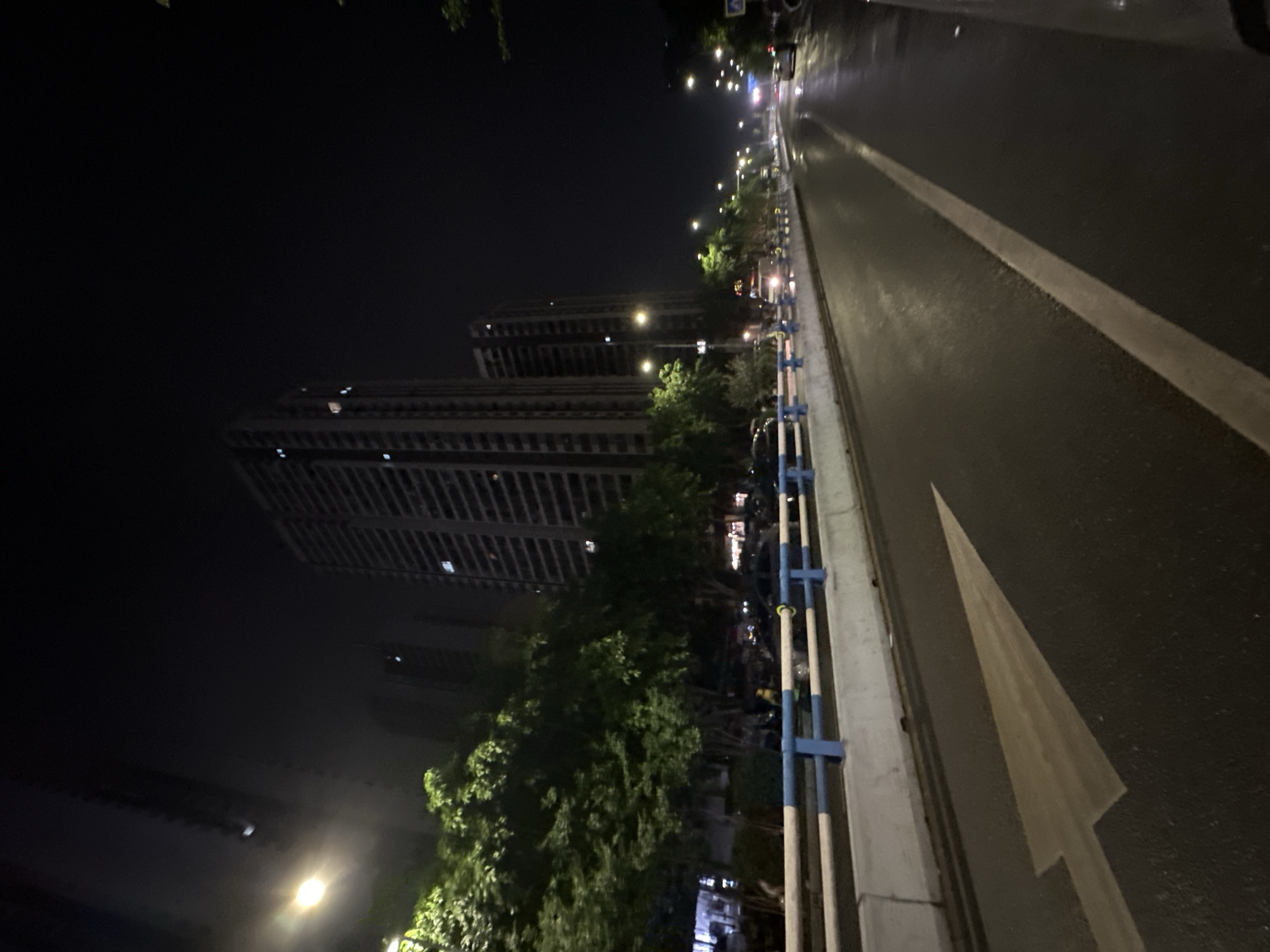 This nighttime image depicts an urban street scene in the Chongqing High-tech Zone, China. The asphalt road in the foreground is visibly wet and highly reflective, indicating recent rainfall. A large, faded white arrow is painted on the road surface, pointing forward and slightly to the left, indicating a straight direction for traffic. A solid white line delineates the right edge of the current lane.
To the left of the road, a concrete median barrier with blue and white horizontal railings separates the lane from a vegetated area. Behind this railing, a dense line of green trees extends into the midground. Partially obscured by the trees and railing, a person wearing a yellow vest or jacket is visible.
Further beyond the trees, on what appears to be a parallel road or service lane, several vehicles, including cars and a white van-like vehicle, are present, some seemingly parked and others potentially in motion. Numerous streetlights, emitting a warm white light, illuminate the scene, casting reflections on the wet road and creating visible halos.
In the background, multiple tall, multi-story buildings, consistent with residential towers, rise into the dark, featureless night sky. The most prominent building on the left exhibits numerous illuminated windows across its facade. Another similar building stands further to the right. A blue and white road sign is faintly visible in the far distance on the right side of the road.