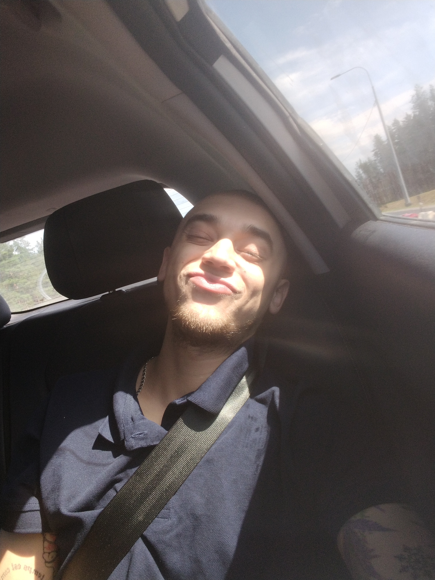 The image shows a young man sitting in the passenger seat of a car, with his eyes closed and a smile on his face. He is wearing a dark blue polo shirt and a silver chain necklace. A seatbelt is across his chest. Sunlight streams in through the car's sunroof, illuminating his face and casting shadows. Outside the car, trees and a street light are visible, suggesting the car is in a suburban or rural area, possibly on the outskirts of a city. Given the user's prompt, this scene could be occurring in Perovo, Russia, where such environments are common. The man appears relaxed and content, enjoying the ride.